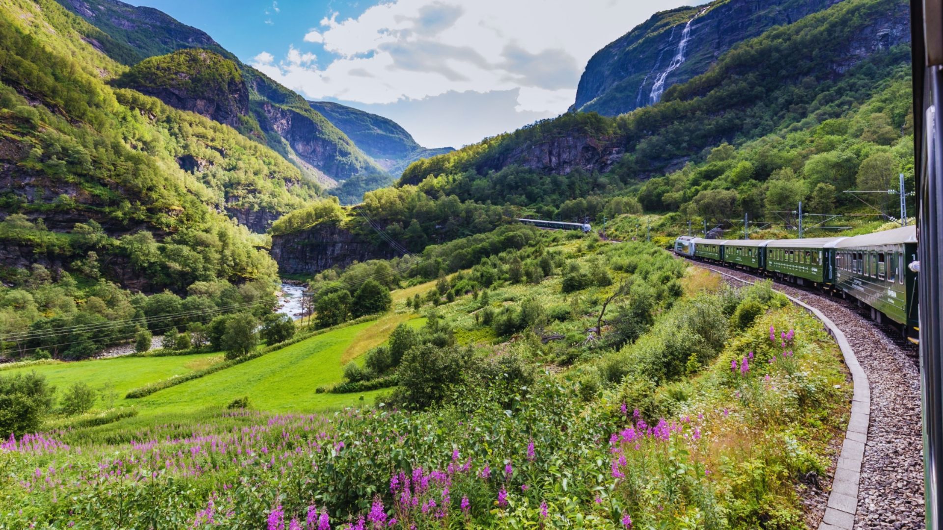 A train moves through a vibrant green valley, surrounded by lush vegetation and rolling hills.