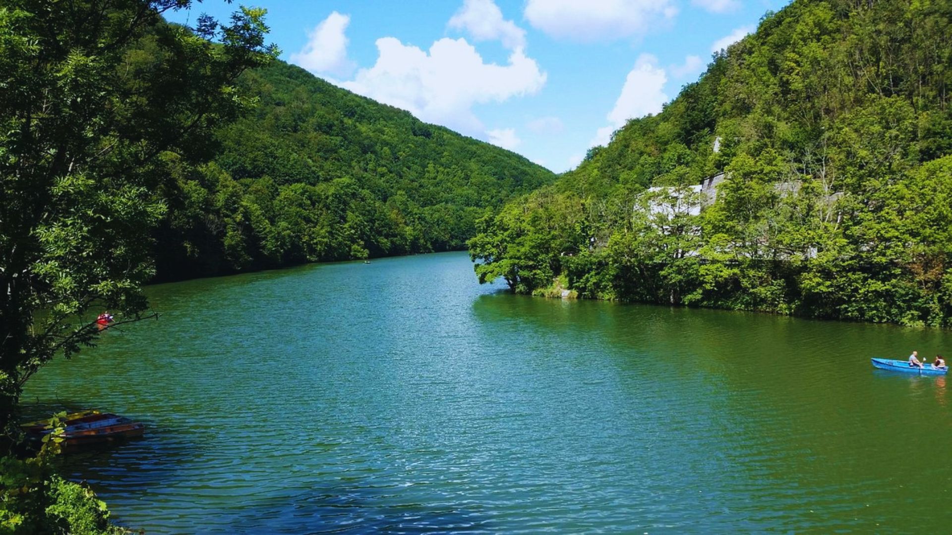 A serene lake bordered by lush, green hills under a clear blue sky. Two people paddle a small blue boat, conveying peace and relaxation.