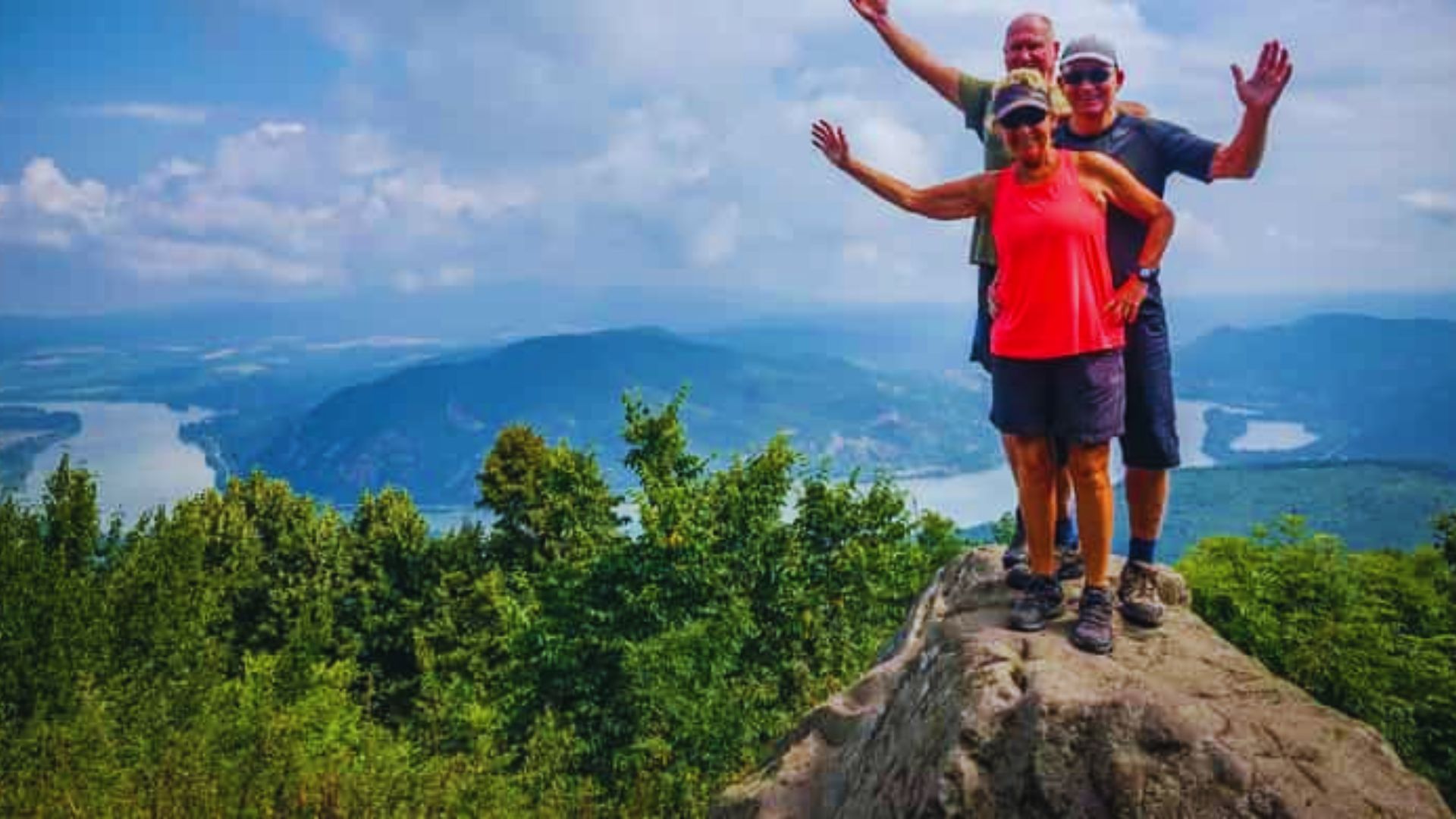 Three people stand triumphantly on a mountain peak, arms raised in celebration against a clear blue sky.