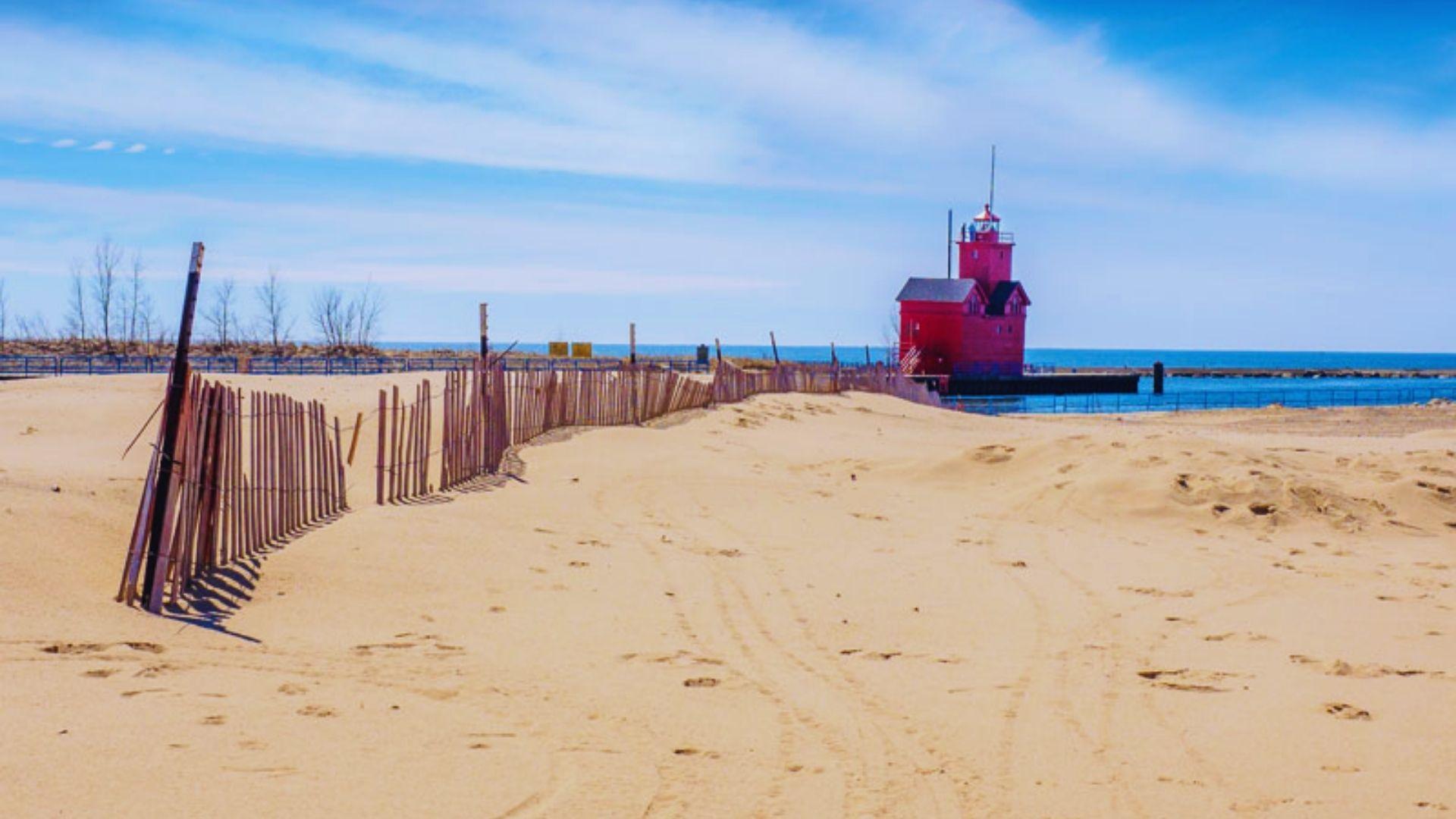 A red lighthouse stands tall on the sandy beach, overlooking the ocean waves under a clear blue sky.