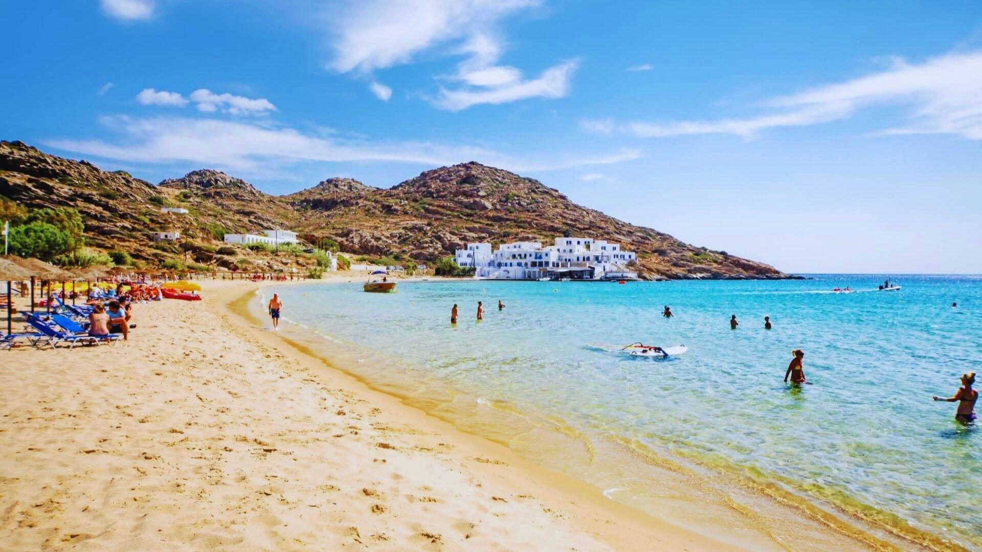A scenic view of Mykonos beach, featuring golden sand, clear blue waters, and sunbathers enjoying the sunny day.