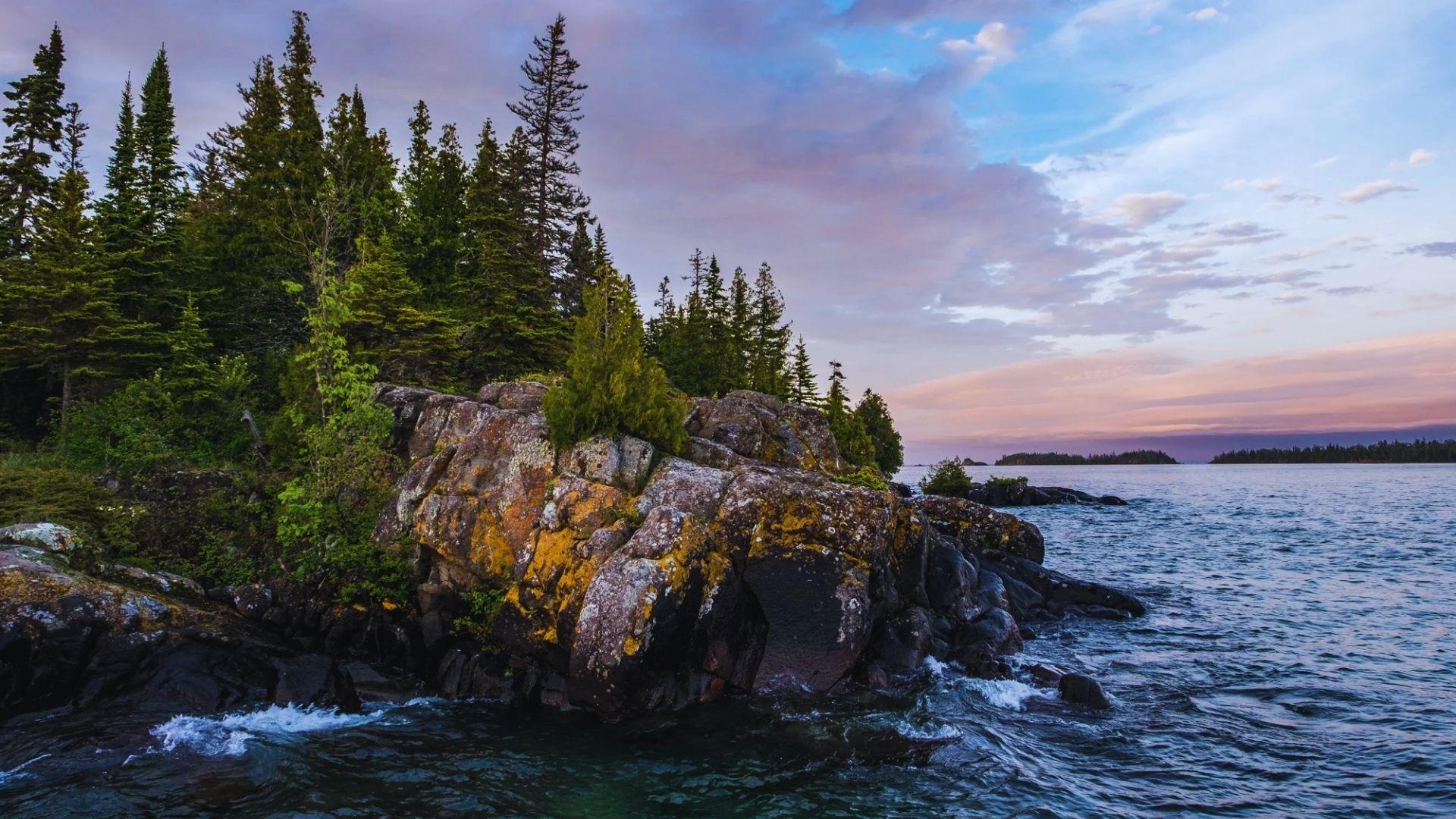 A rocky shoreline with silhouetted trees and calm water, illuminated by a vibrant sunset sky.