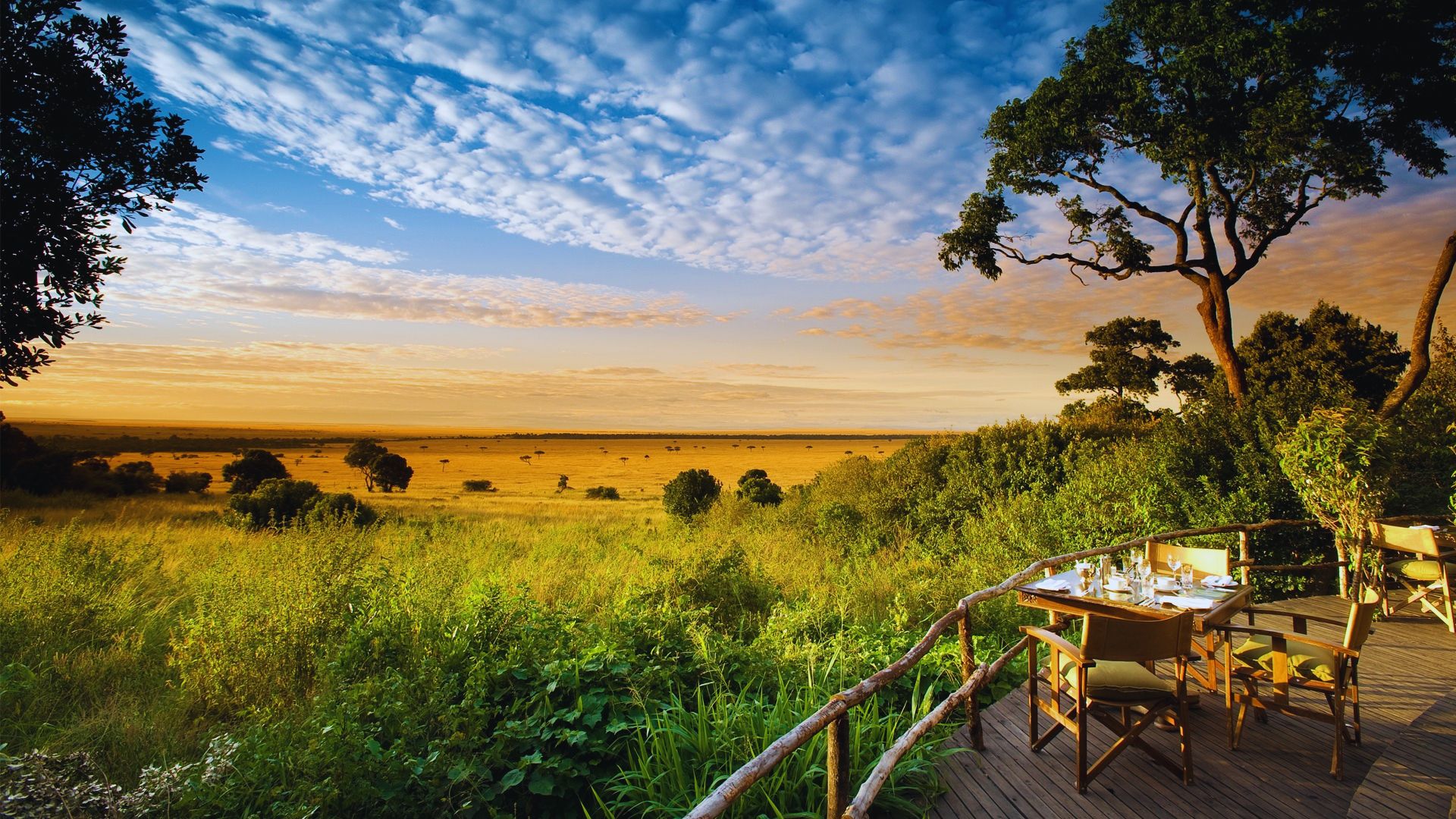 The sun sets over the savannah, painting the sky with orange and pink from a deck perspective.