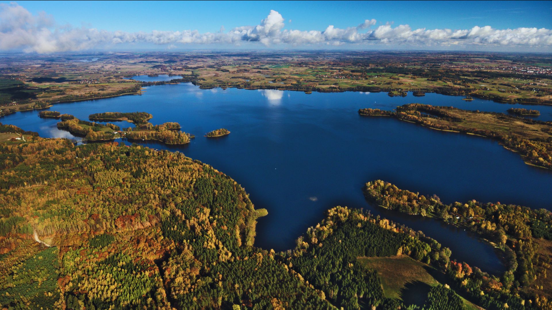 Aerial view of a vast blue lake surrounded by lush green and autumn-colored forests under a clear blue sky with scattered clouds, conveying tranquility.