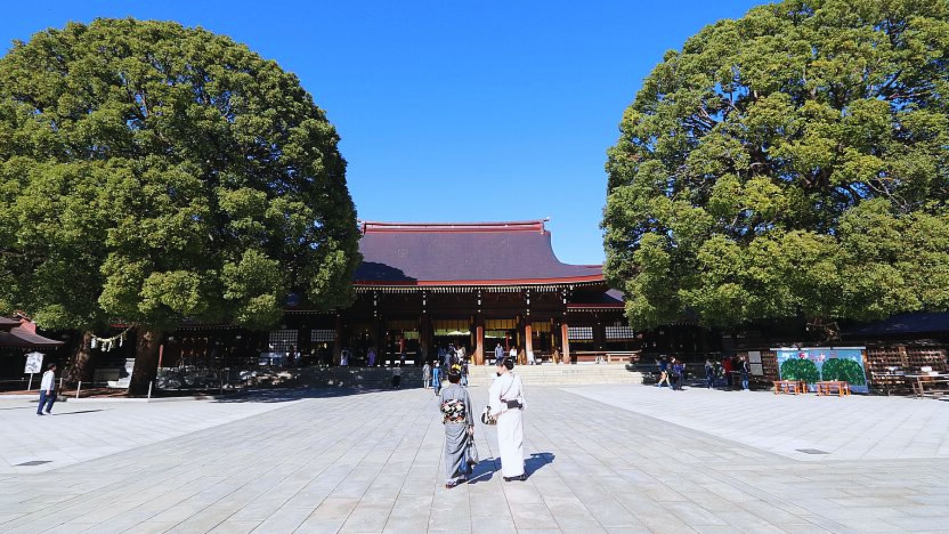 Two individuals stand in front of a large building, showcasing its impressive architecture and scale.