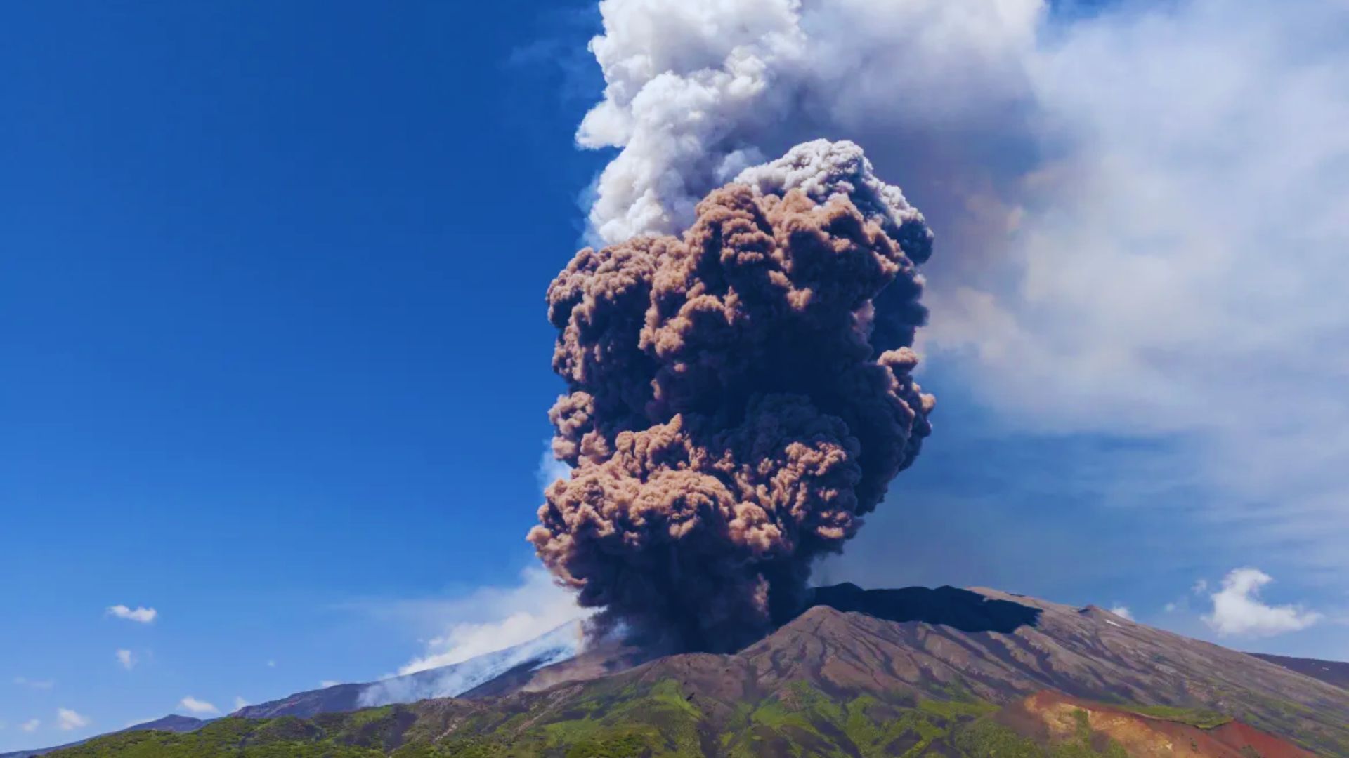 A large plume of smoke rises from the summit of a mountain, indicating possible volcanic activity or a fire.