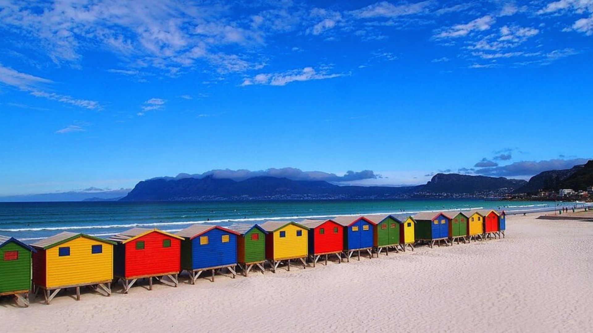 Colorful beach huts are lined up along the shore, with a majestic mountain rising in the background.