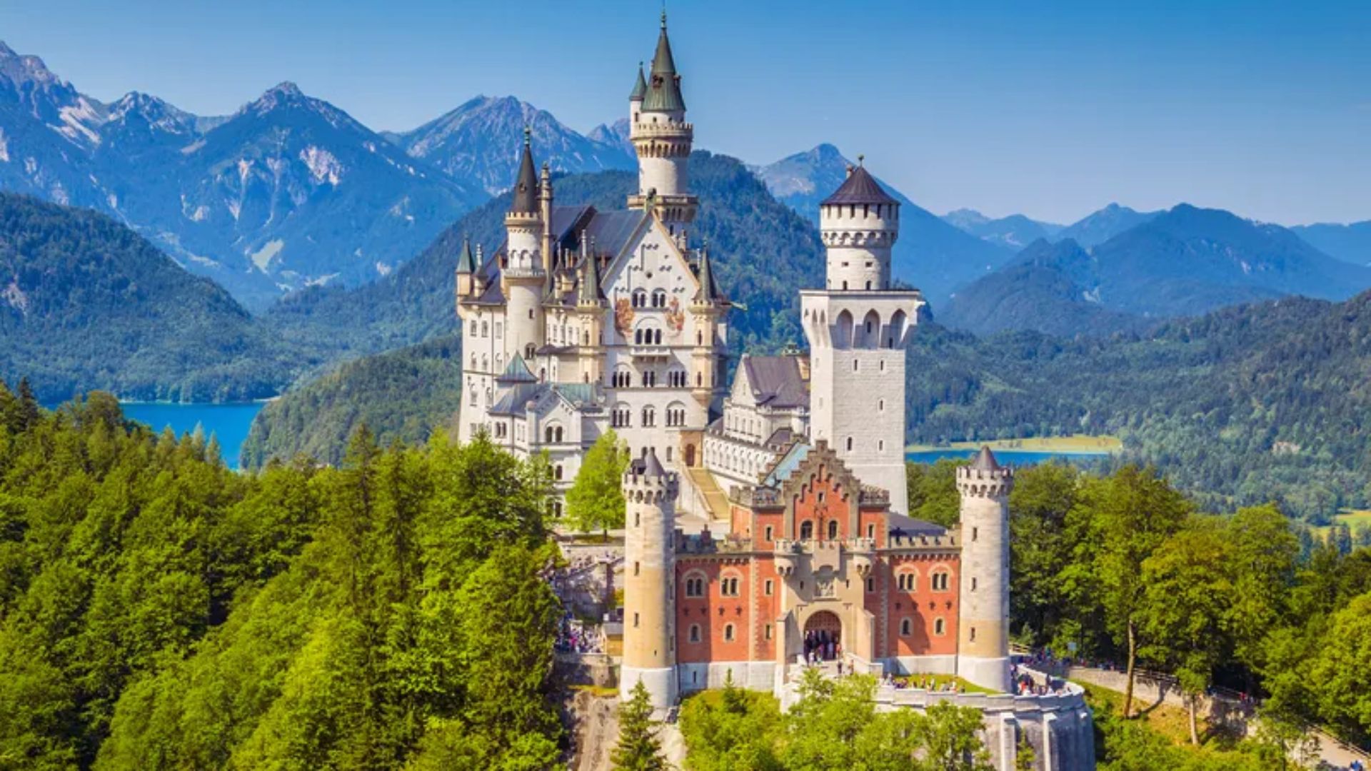 Neuschwanstein Castle in Germany, a fairy-tale structure surrounded by lush forests and mountains under a clear blue sky.