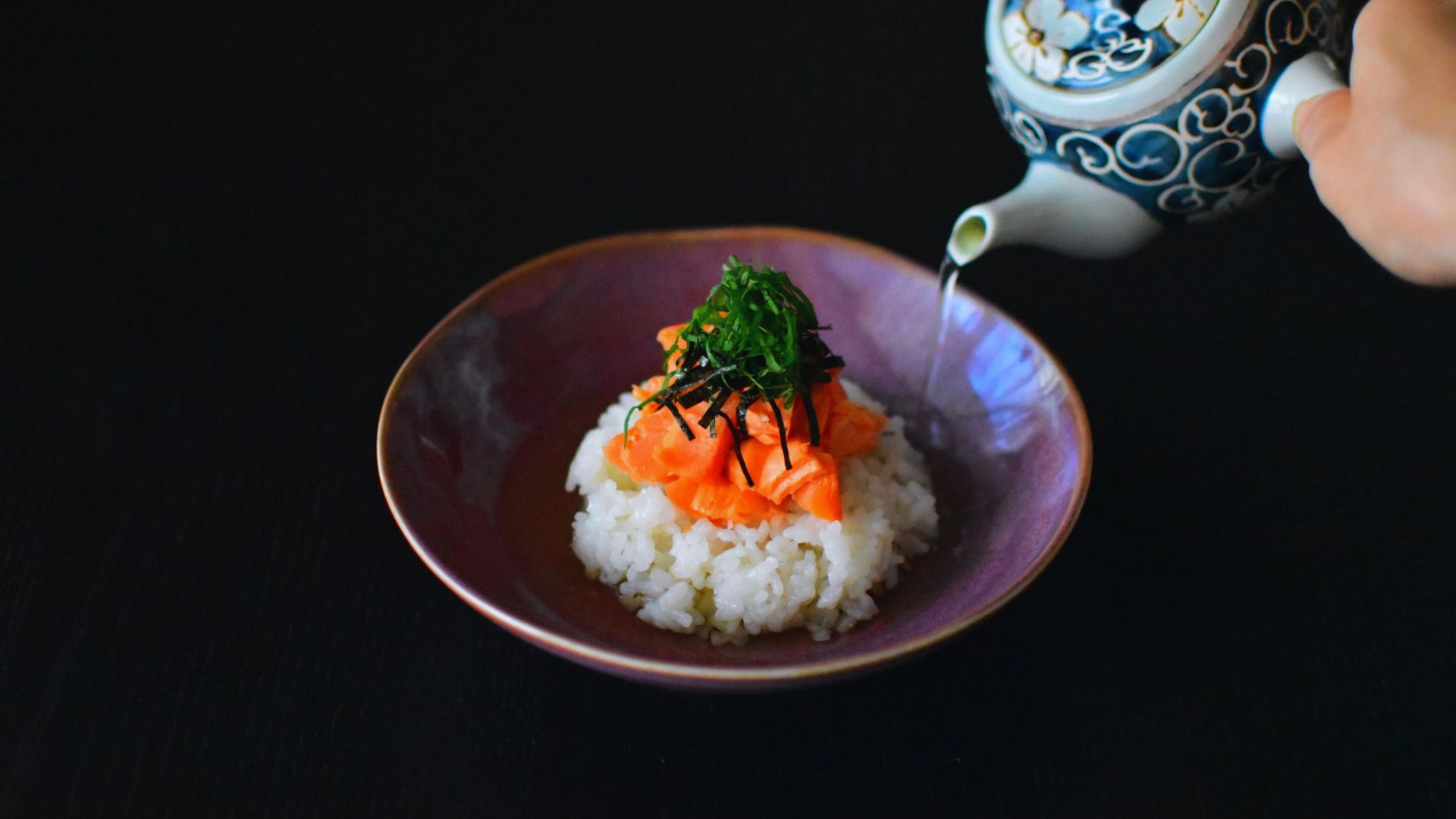 A person is pouring tea from a teapot onto rice and salmon, adding a flavorful touch to the meal.