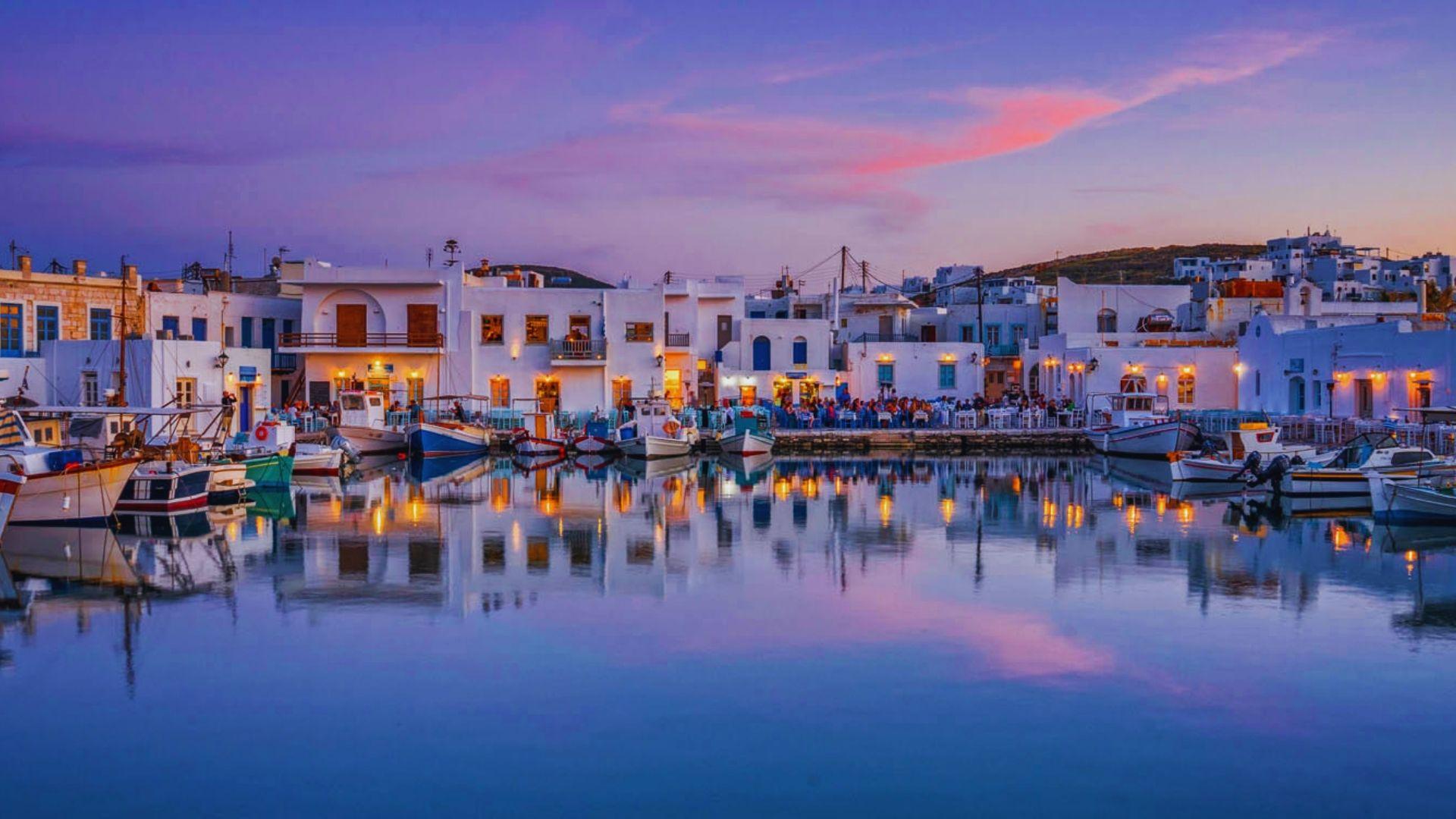 Sunset over the port of Mykonos, Greece, with vibrant colors reflecting on the water and silhouettes of boats.