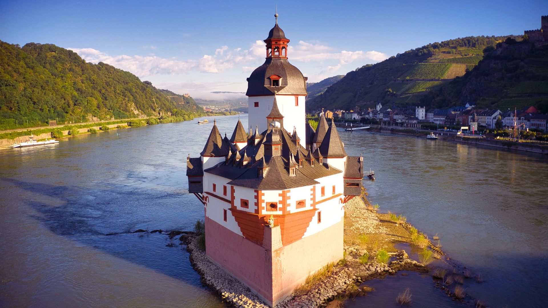. A majestic castle perched on a hill overlooks a serene river below, surrounded by lush greenery.
