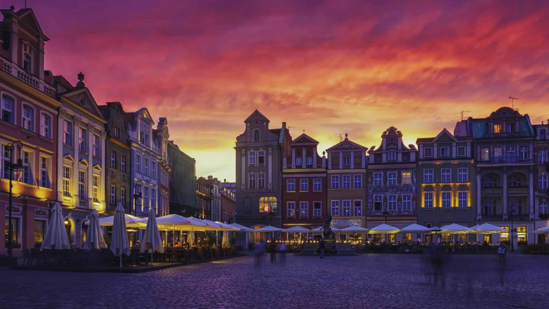 Colorful historic buildings line a plaza under a dramatic sunset sky, with deep purples and oranges. Umbrellas and blurred figures evoke a lively atmosphere.
