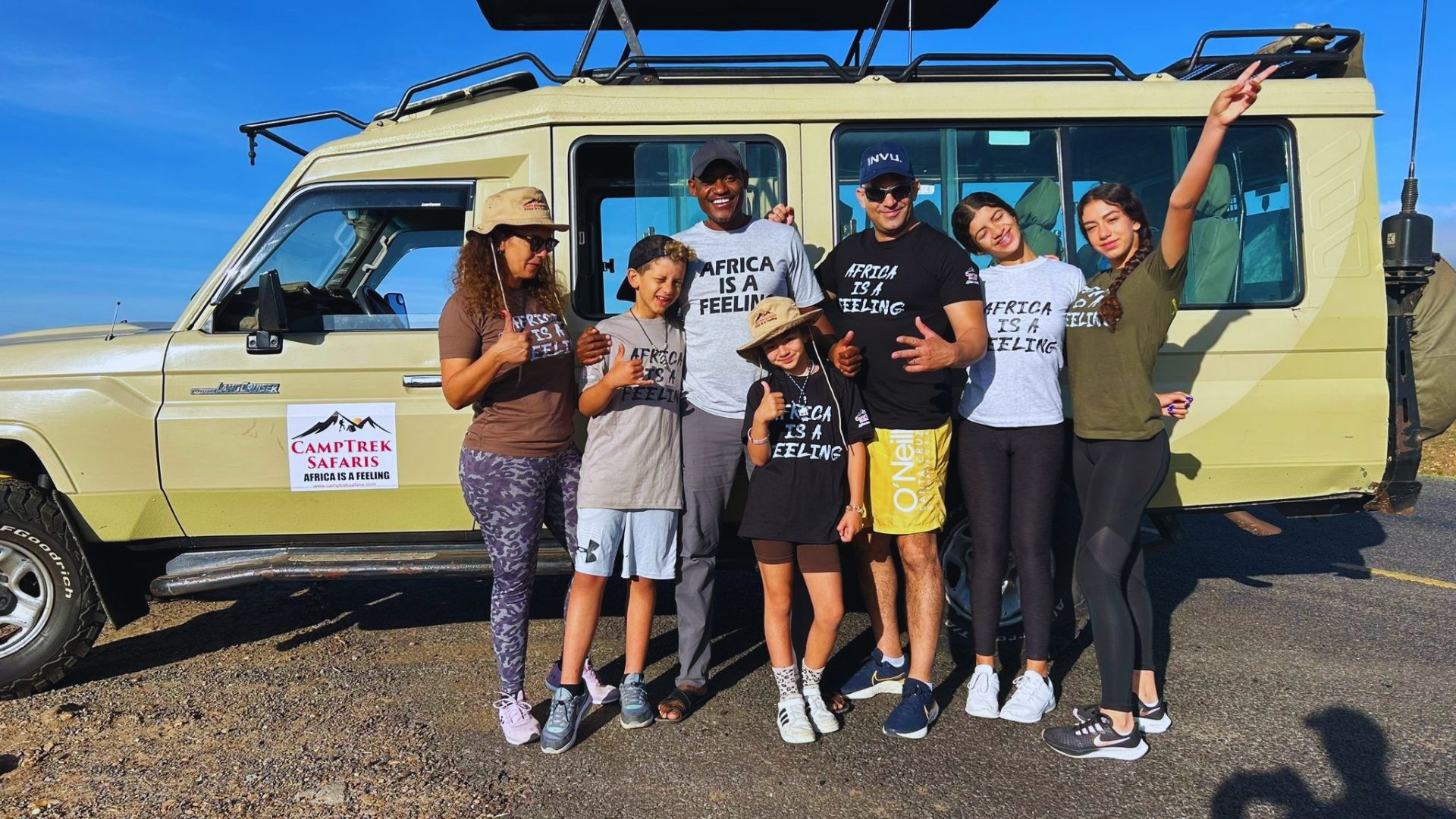A family smiles together for a photo in front of their parked vehicle on a sunny day.