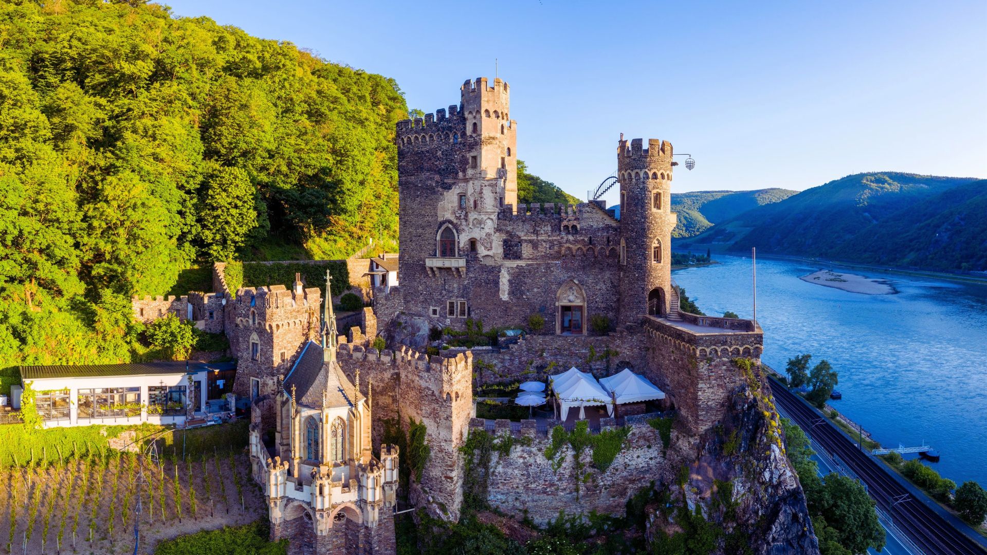 A castle sits atop a hill, overlooking a serene river below, surrounded by lush greenery.