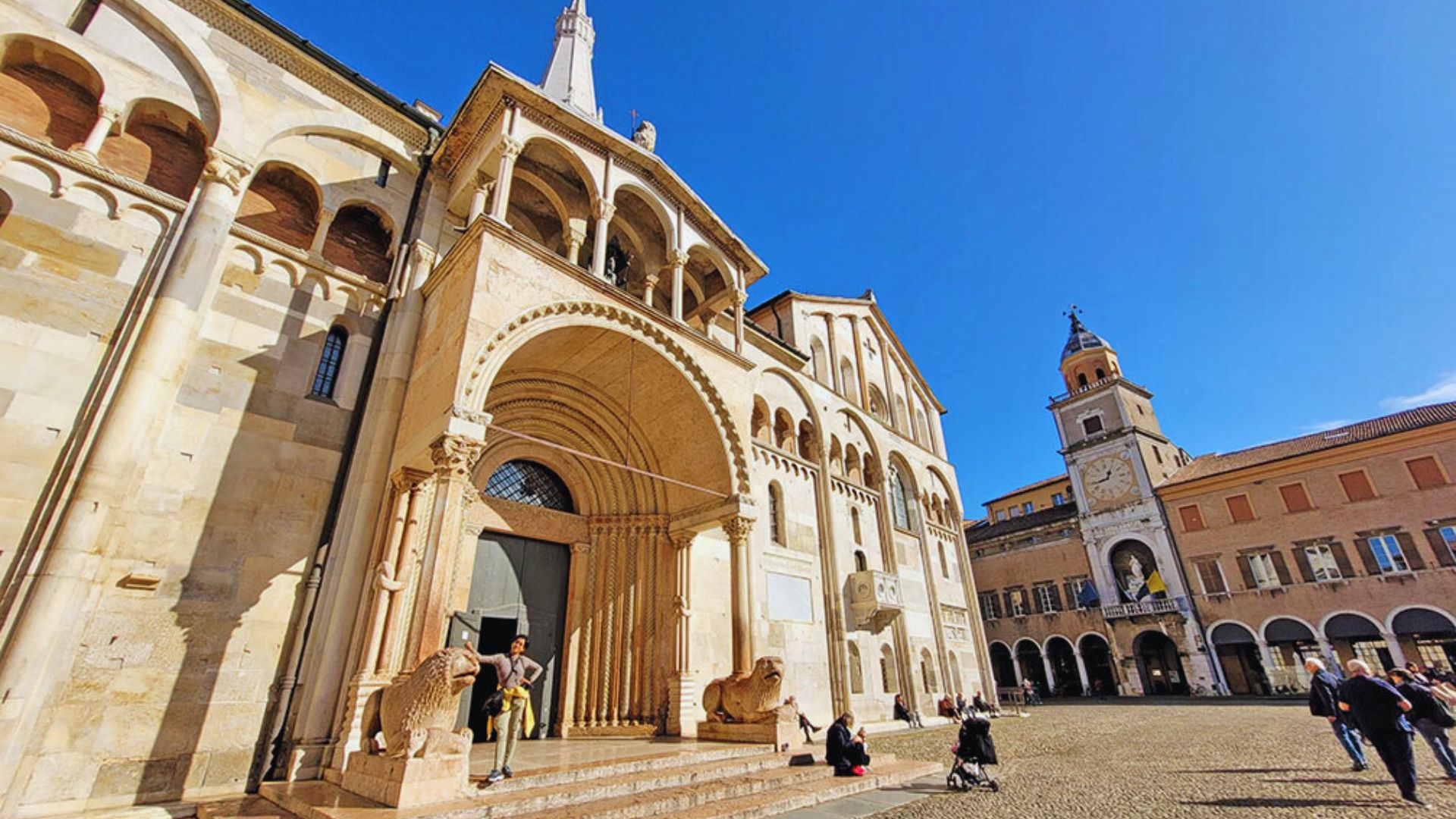 Historic Italian plaza with grand Romanesque cathedral entrance, elaborate arches, and stone lions under a clear blue sky; people walking nearby.