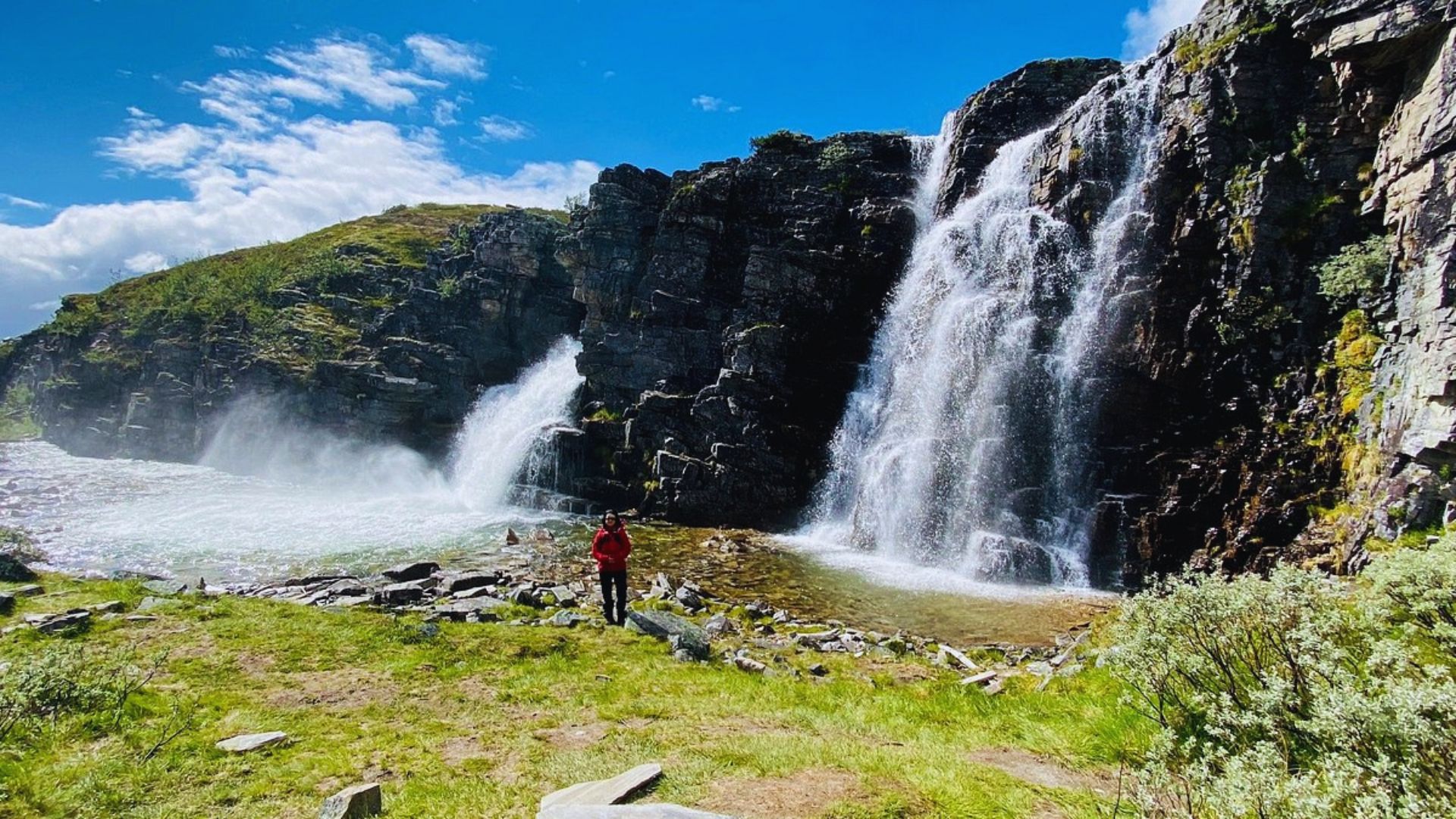 A person stands in front of a cascading waterfall, surrounded by lush greenery and mist rising from the water.