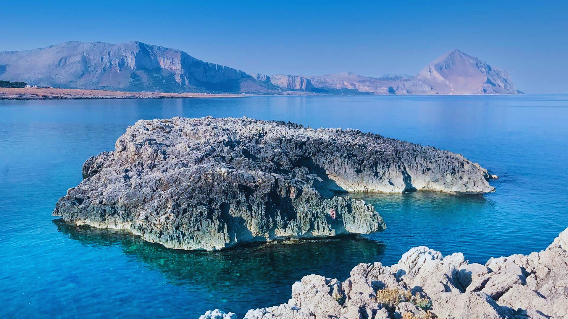 A rugged rocky outcrop rises from the ocean, surrounded by waves and blue water under a clear sky.