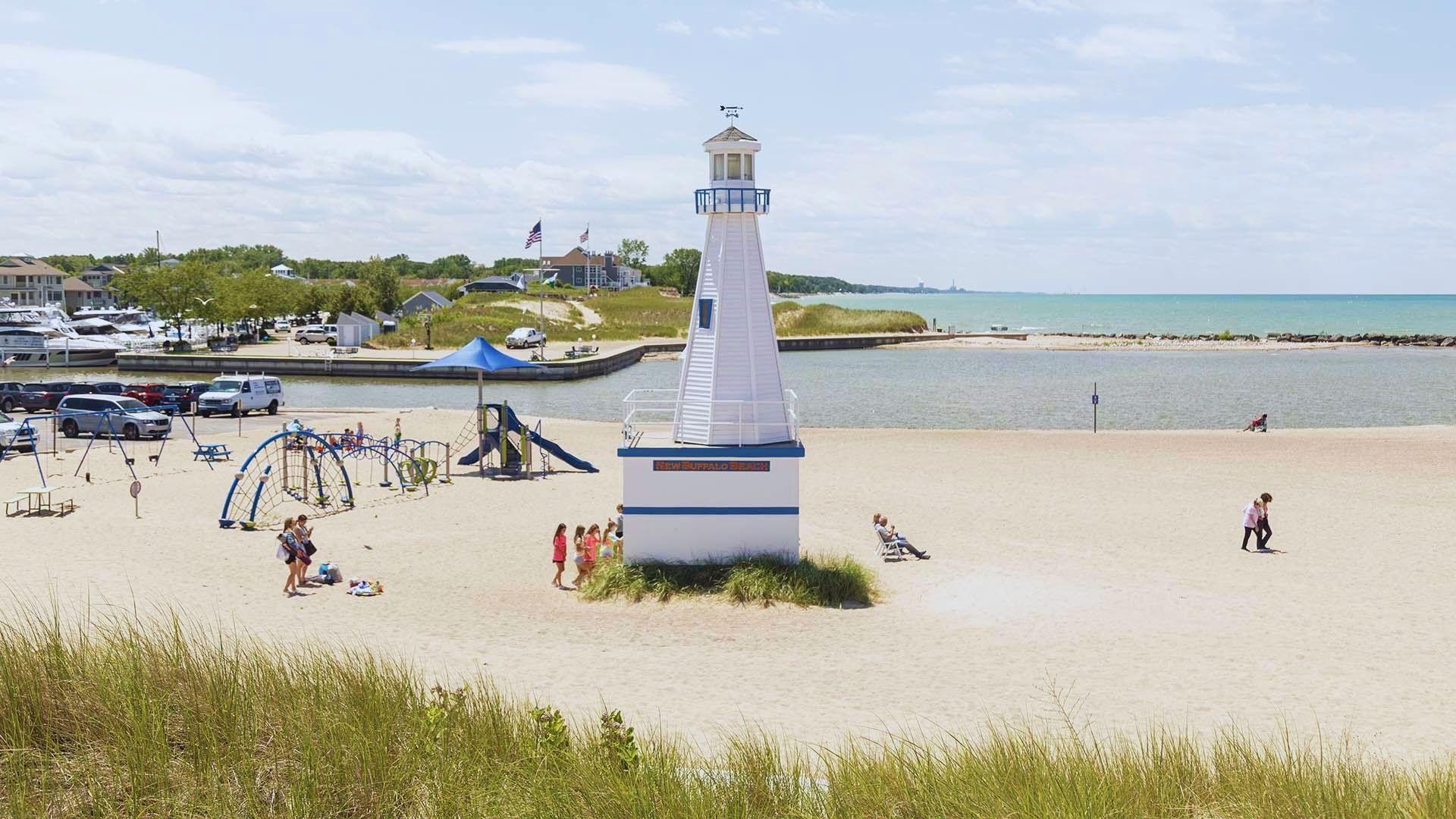 A lighthouse stands on the beach as people enjoy playing in the sand nearby under a clear blue sky.