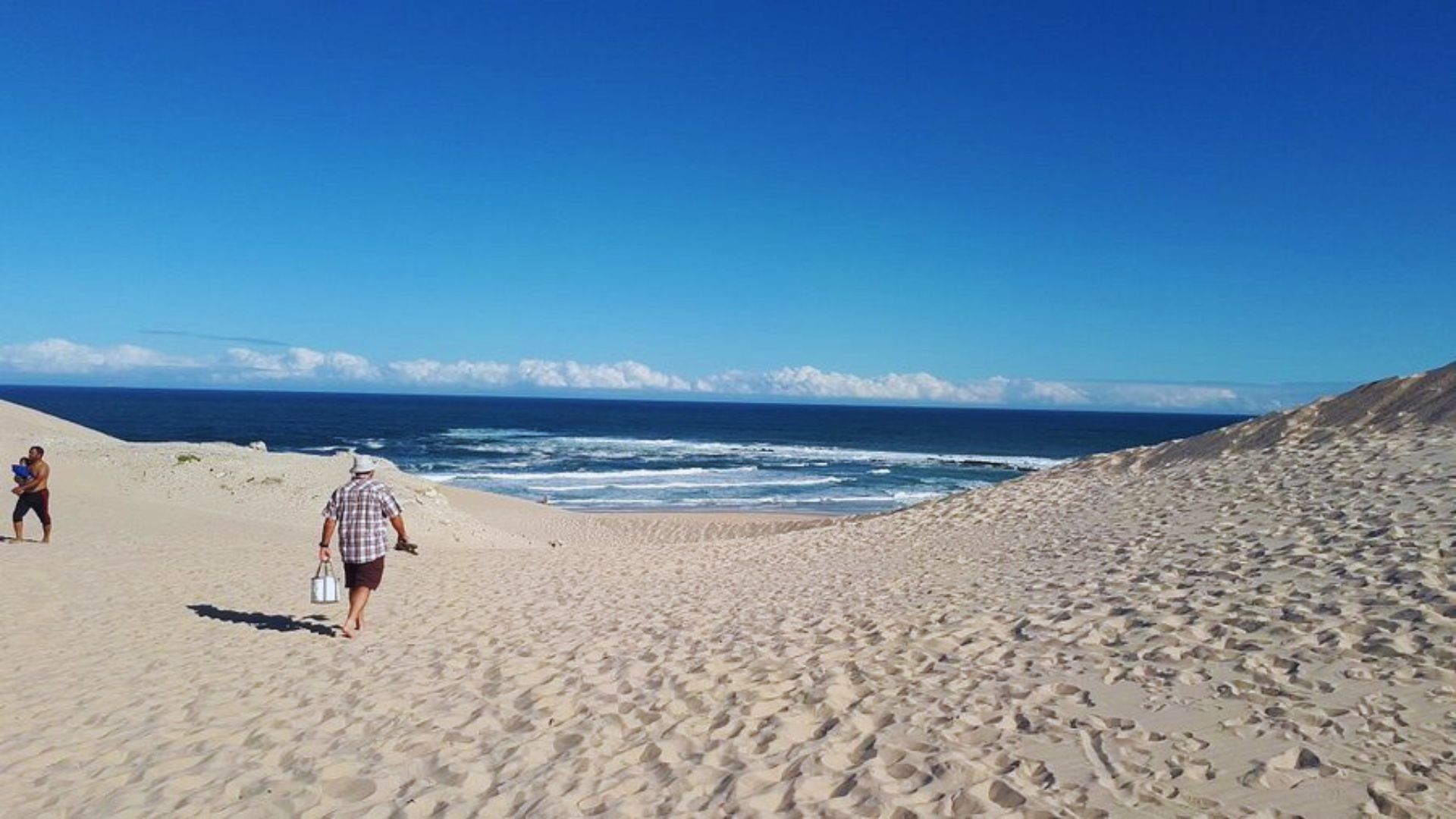 People stroll along a sandy beach, with gentle ocean waves lapping at the shore in the background.