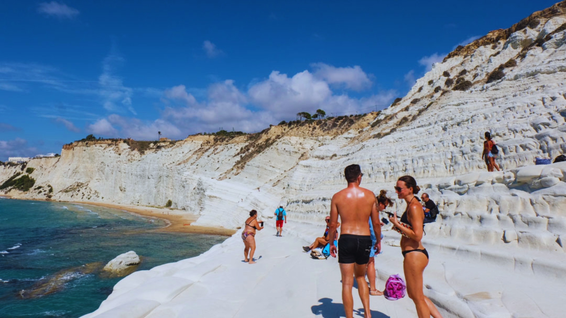 People stroll along the beach, with striking white cliffs rising in the background under a clear blue sky.