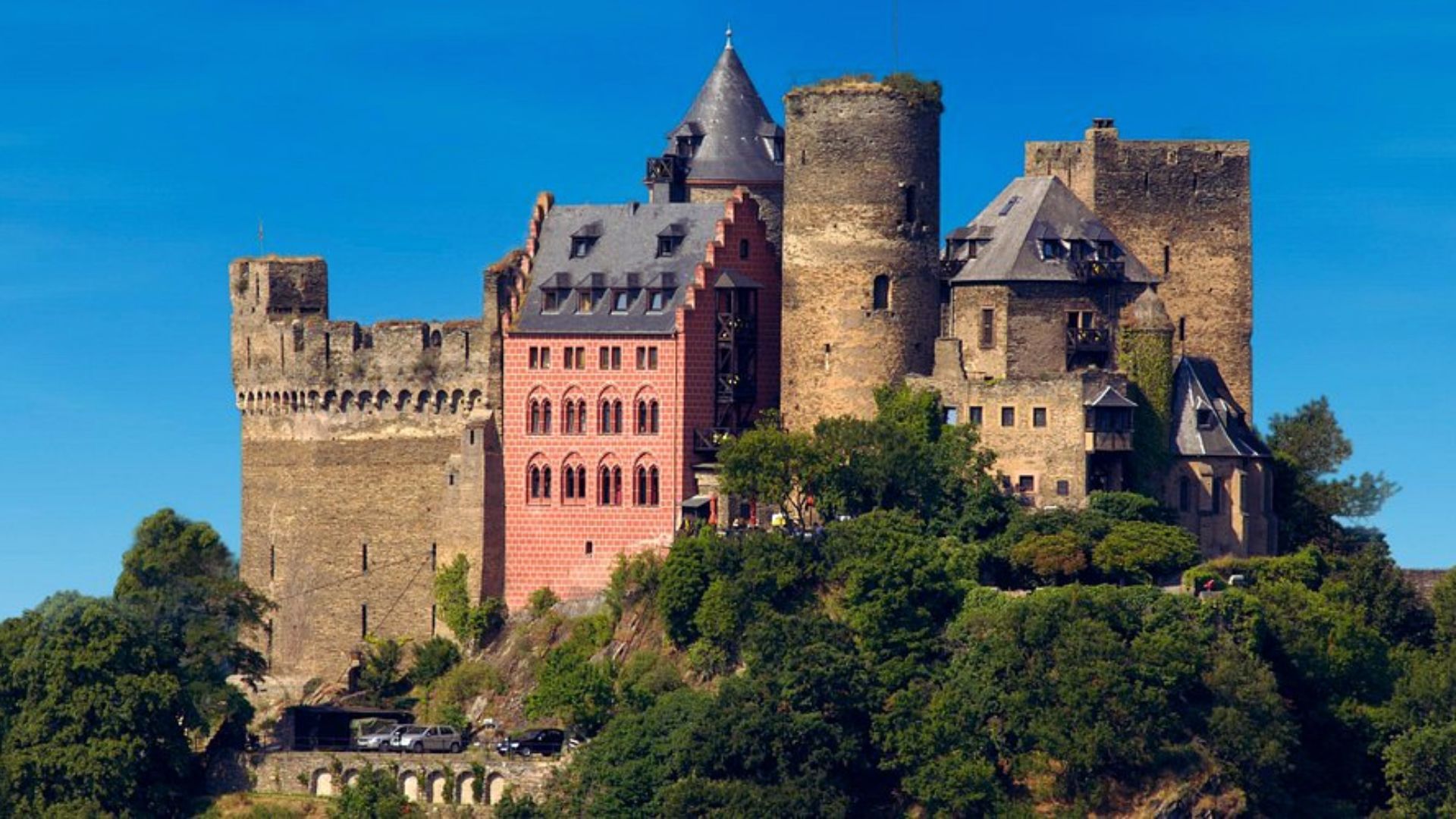 A scenic view of a historic castle overlooking the Rhine River in Germany, surrounded by lush greenery and blue skies.