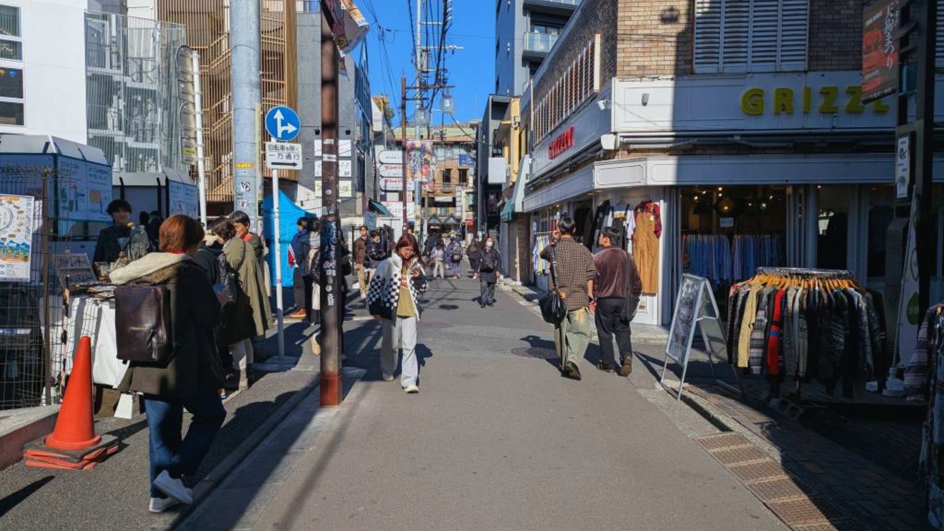 People walking along a narrow street in an Asian city, surrounded by traditional buildings and vibrant street life.