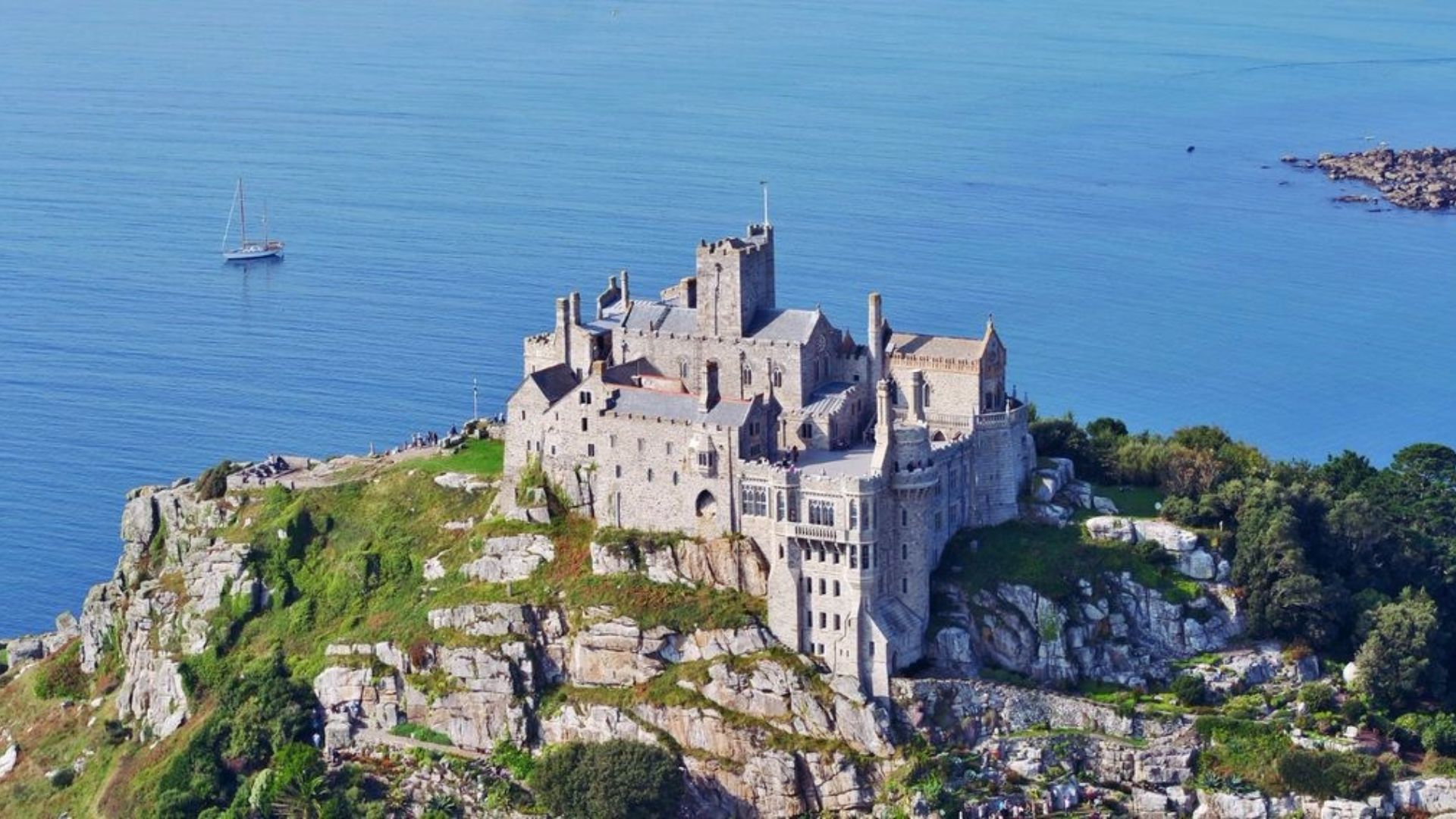 Aerial view of a majestic castle perched atop a green hill, surrounded by trees and open landscape.
