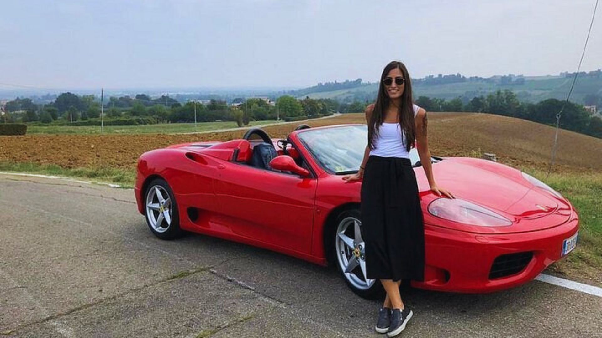 A woman stands next to a sleek red sports car, exuding confidence in a casual outfit on a clear day.