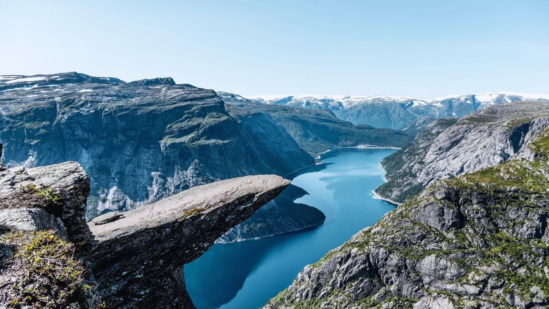 Scenic view from a cliff edge overlooking a tranquil lake surrounded by lush greenery and distant mountains.