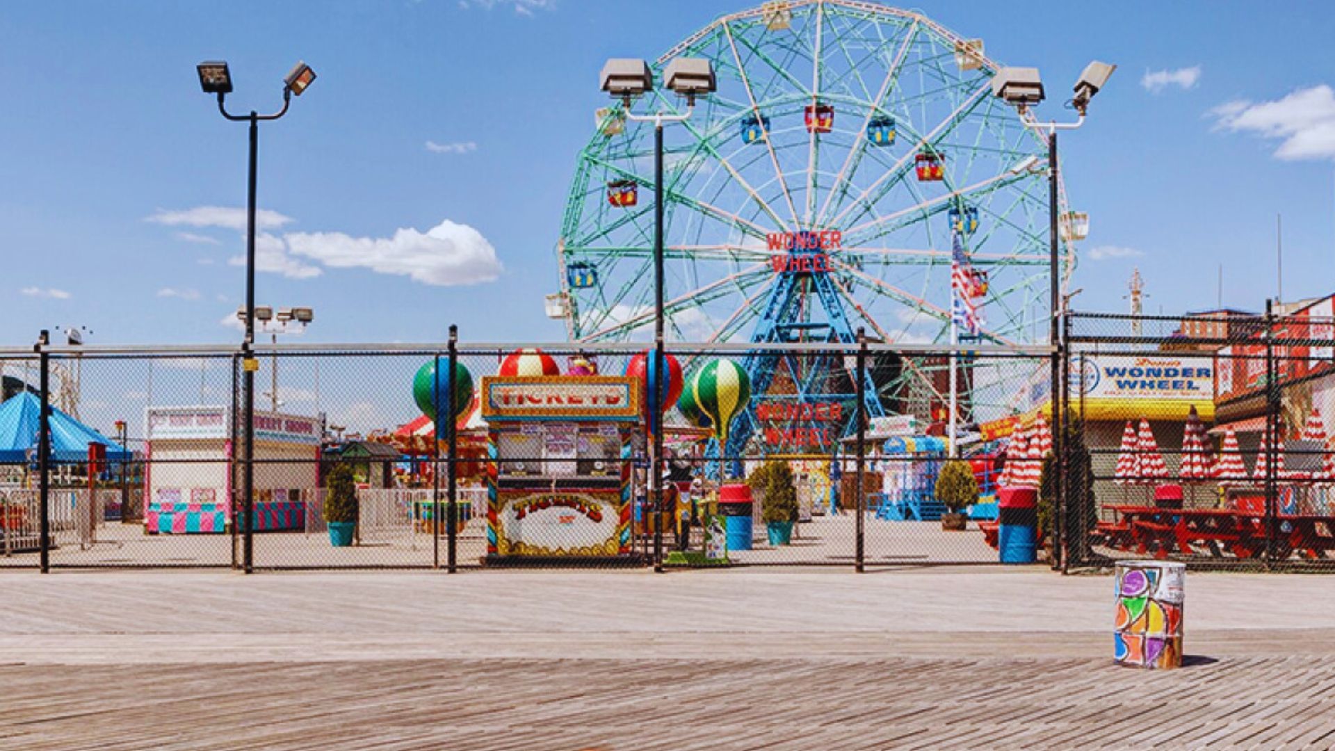 An amusement park scene featuring a ferris wheel and rides, set against a backdrop of a wooden fence.