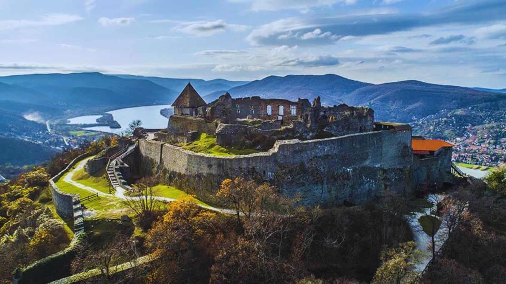 Aerial view of a medieval hilltop castle ruins surrounded by lush forests and winding river. Rolling hills in the background under a partly cloudy sky.