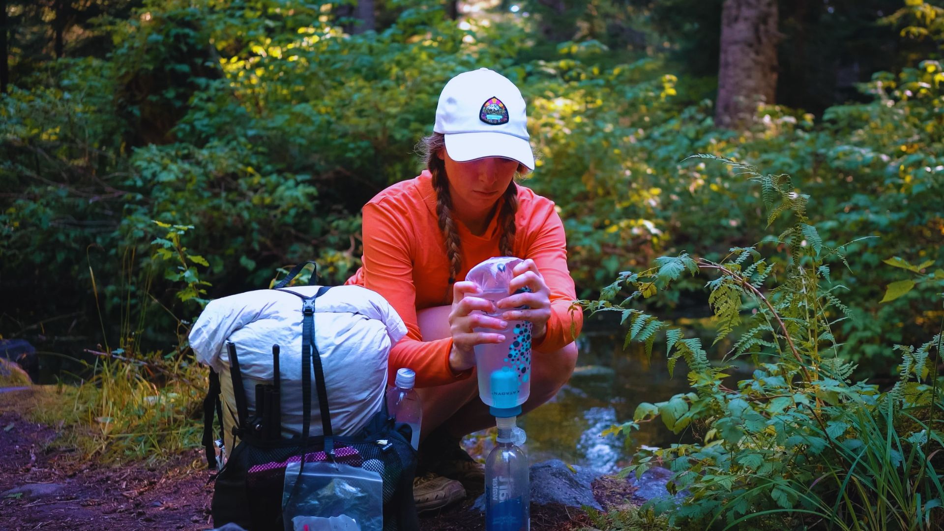 A woman wearing an orange shirt and hat sits beside a stream, enjoying the peaceful natural surroundings.