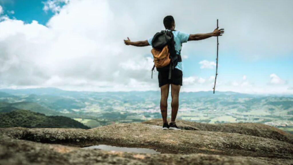 What Is the Safest Way to Travel? Full Travel Guide A man with a backpack stands on a mountain summit, arms raised in triumph, enjoying the expansive view below.