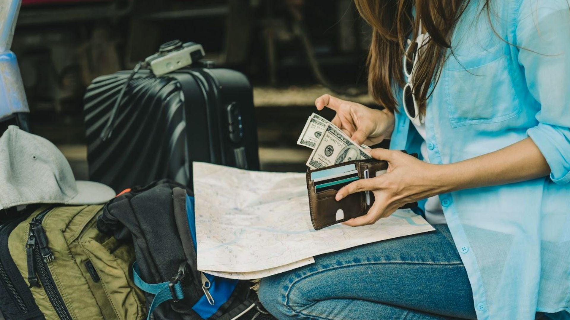 A woman sitting on the ground beside her luggage, looking contemplative in an outdoor setting.