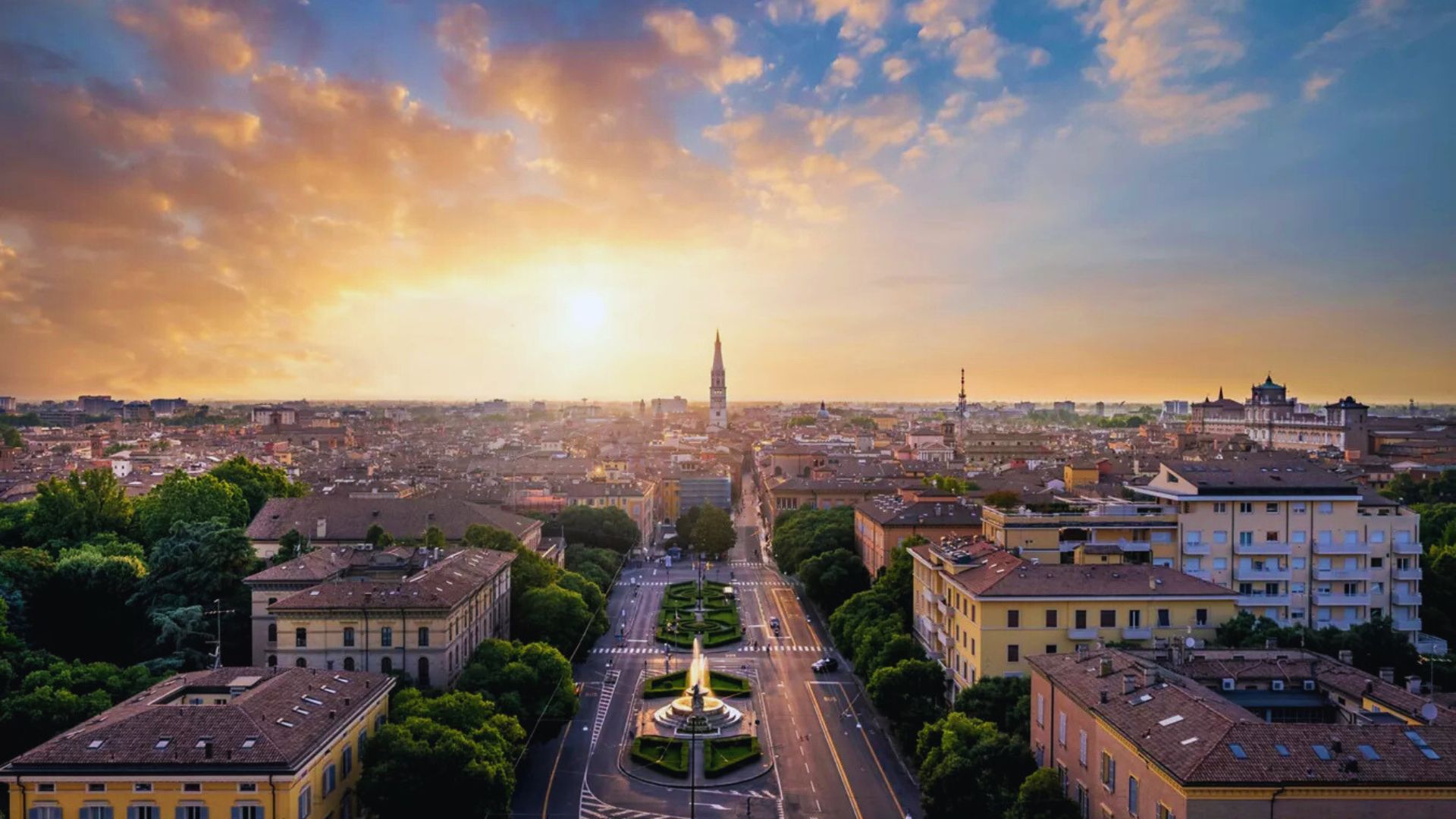 Aerial view of a cityscape at sunset, with a prominent central road lined by trees leading to a distant tower. Soft sunlight and scattered clouds create a serene atmosphere.