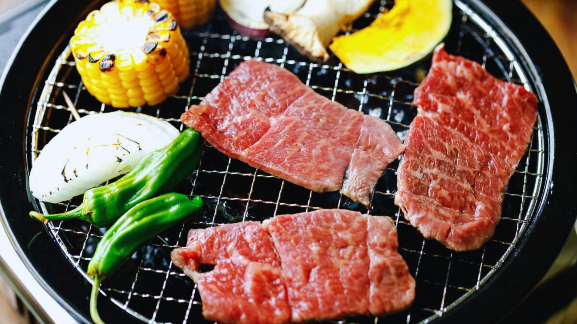 Grilled beef steak alongside corn and assorted vegetables on a barbecue grill.