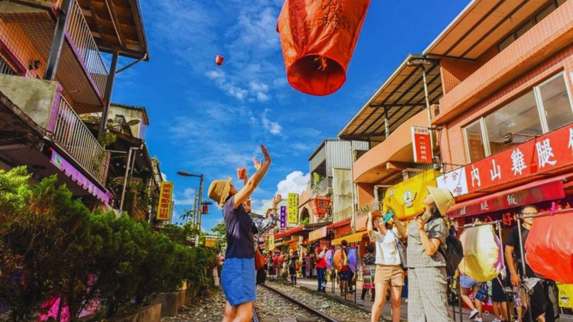 People release colorful lanterns into a bright blue sky on a lively street with vibrant buildings. The scene conveys a festive and joyful atmosphere.