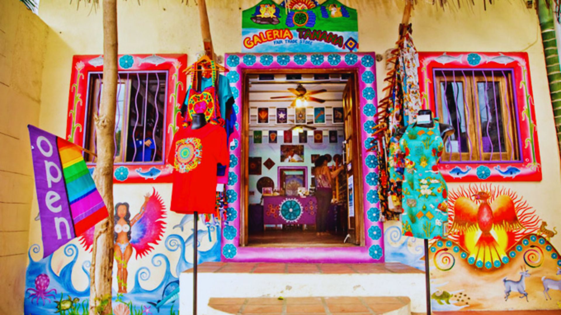 Colorful art gallery storefront with vibrant murals, clothing displays, and an "open" flag. The entrance is adorned with bright decorative motifs.