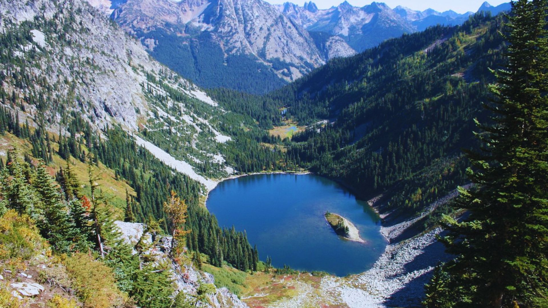 A serene mountain lake surrounded by dense forests and towering peaks under a clear blue sky. The scene conveys tranquility and natural beauty.