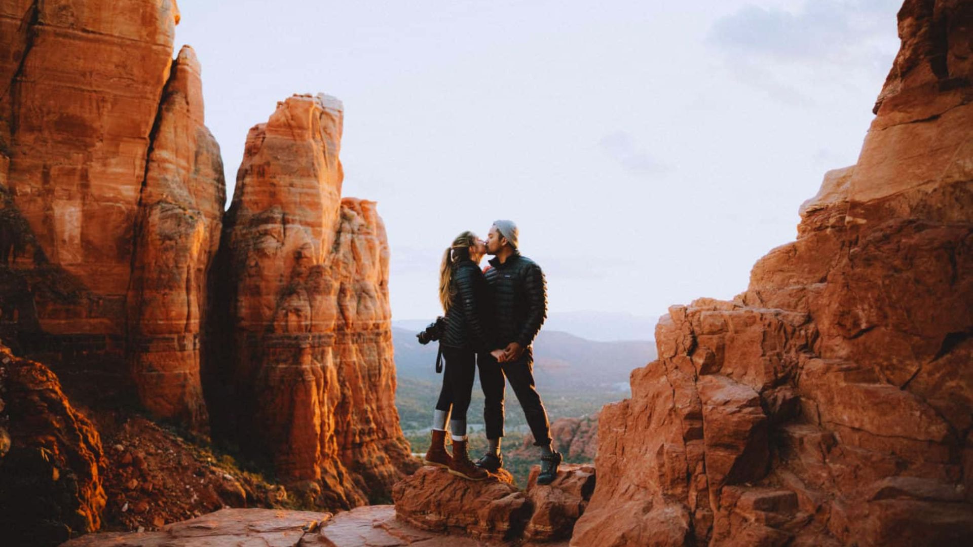 A couple kisses on a rocky ledge with towering red rock formations and a vast landscape in the background, creating a romantic and adventurous mood.