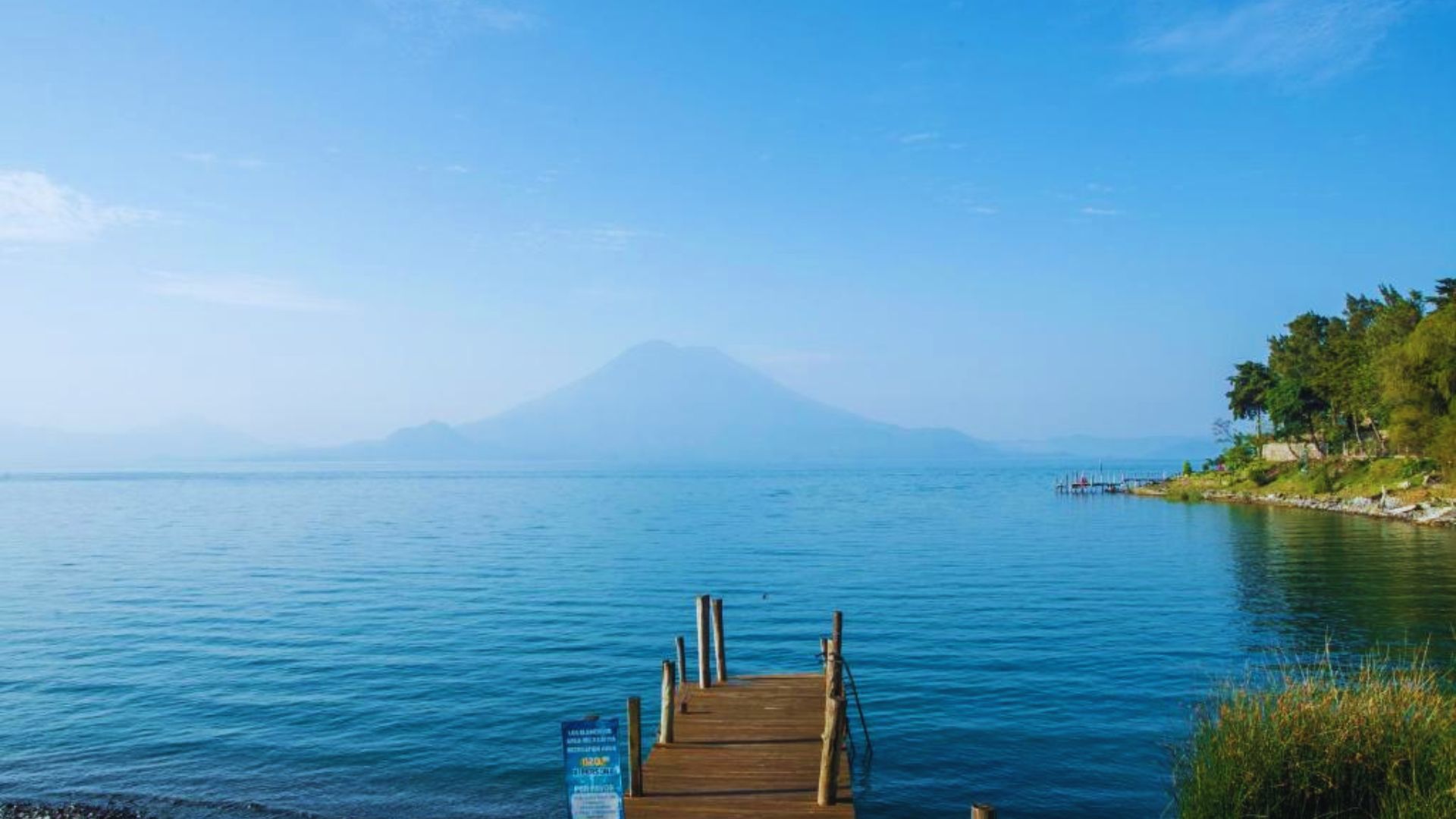 A wooden dock extends over calm water, with a majestic mountain rising in the background under a clear blue sky.