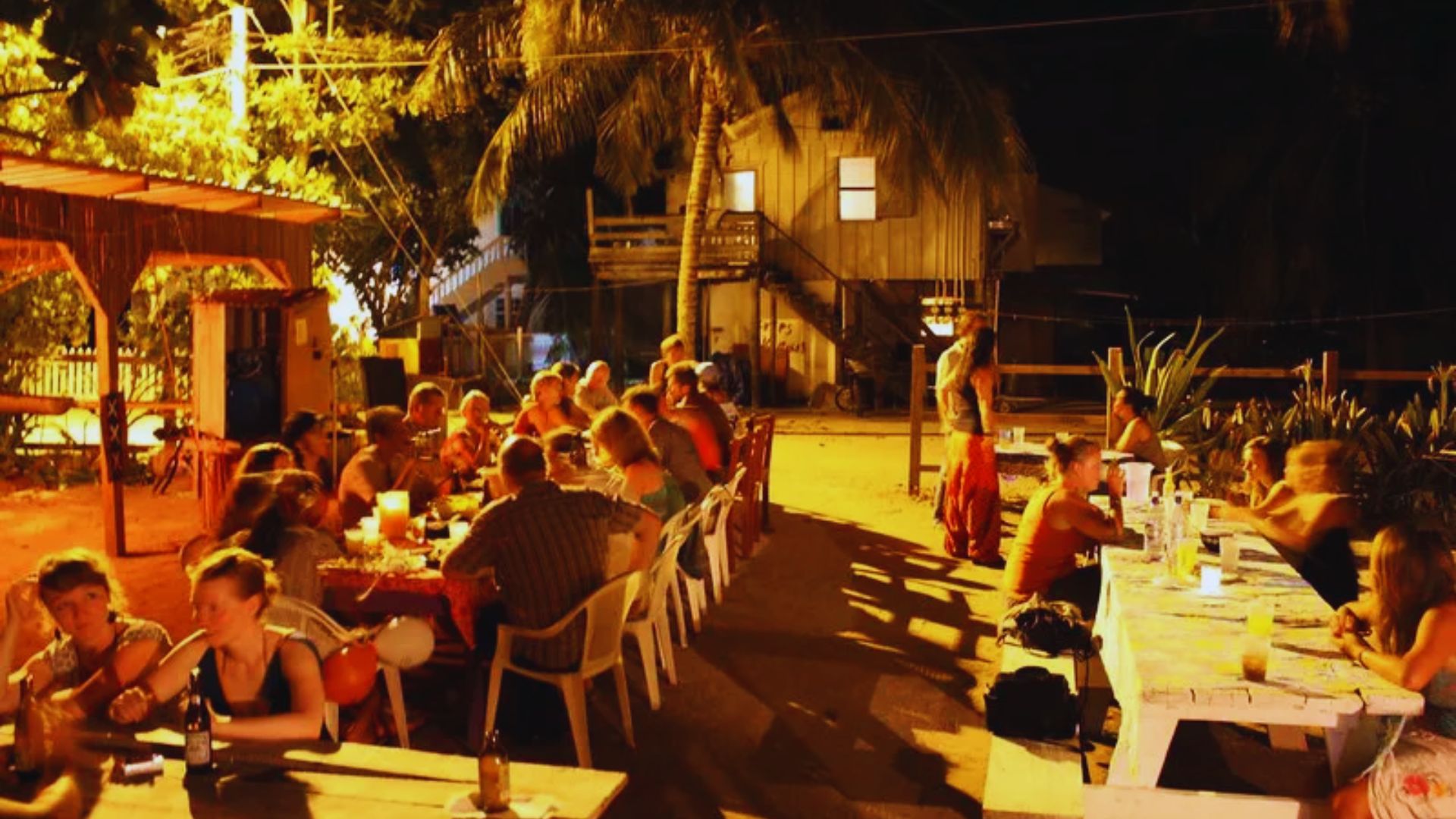 A group of people gathered around tables at night, engaged in conversation under soft lighting.