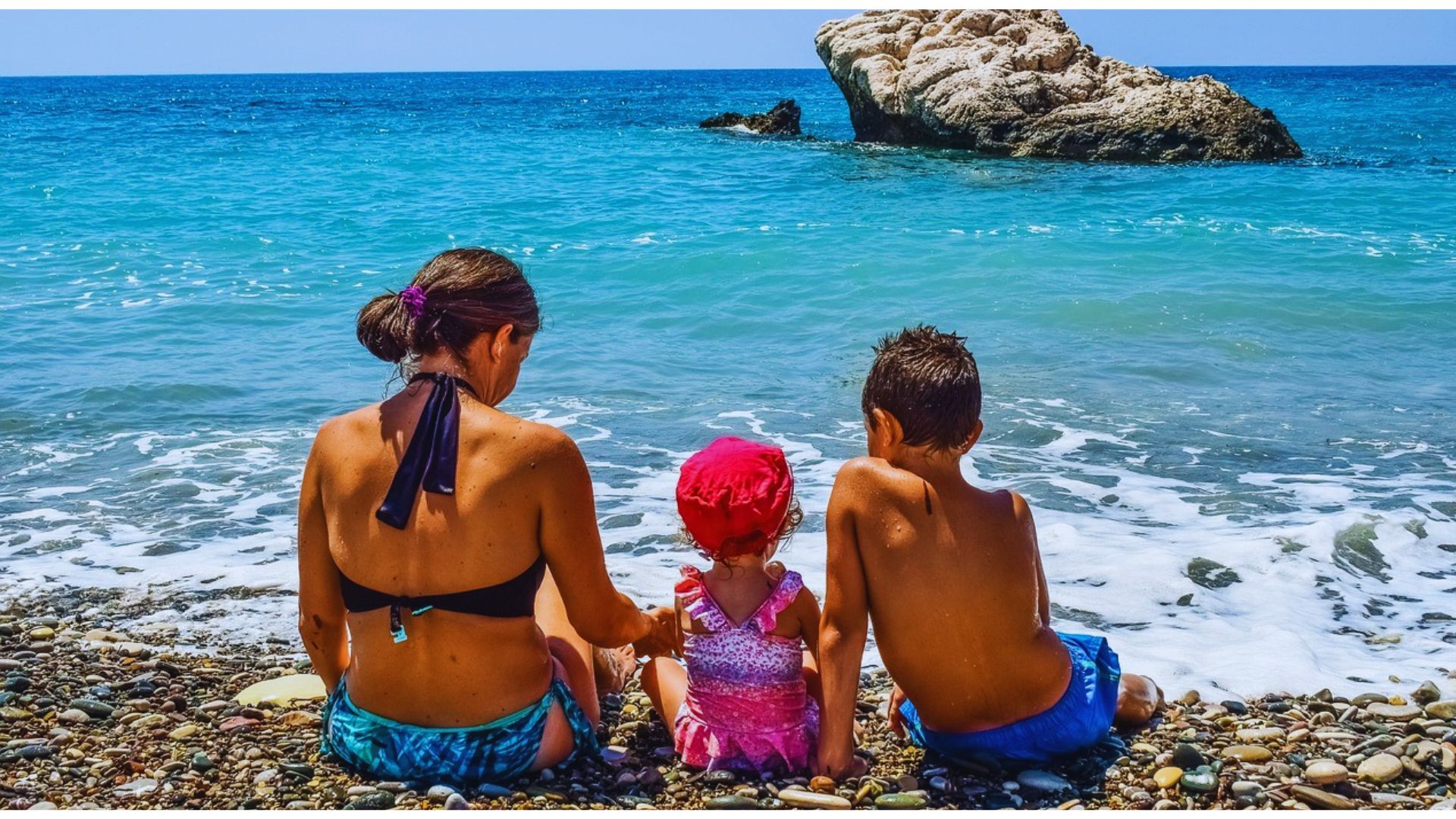 A family sits on a pebbled beach, facing the calm blue sea. A mother and two children relax together, creating a peaceful and content atmosphere.