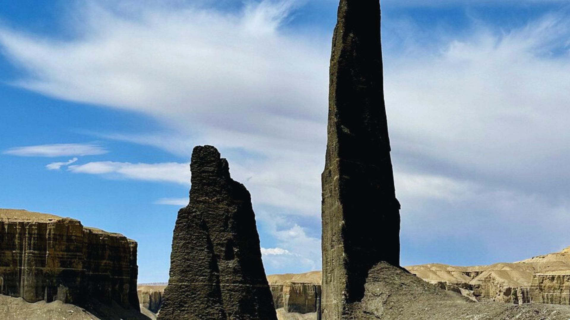A towering, narrow rock formation stands against a deep blue sky with wispy clouds. It is set among desert cliffs, conveying a sense of vastness and awe.