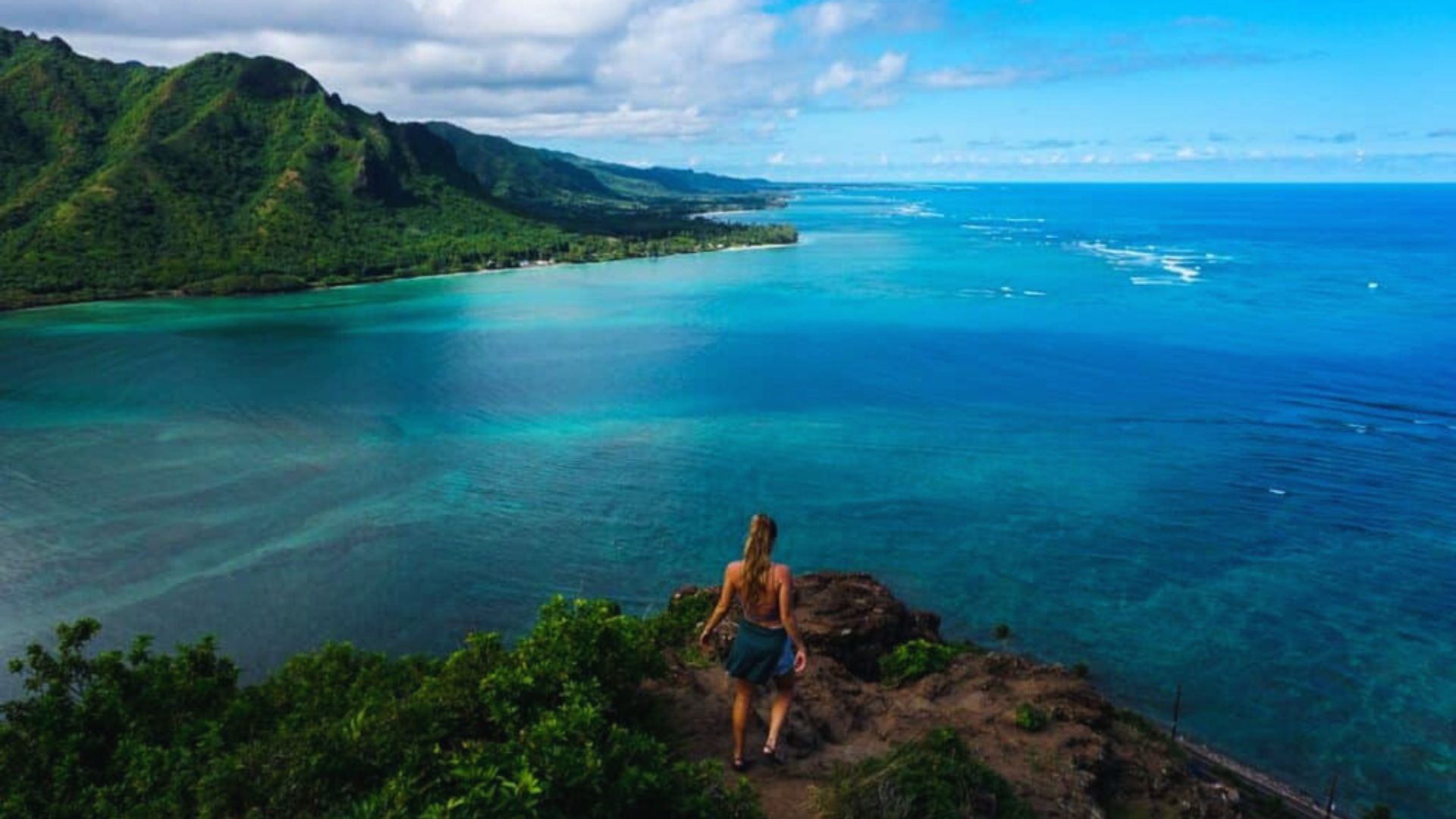 A woman stands on a cliff overlooking a vast, turquoise ocean with lush green mountains in the distance under a partly cloudy sky, conveying tranquility.