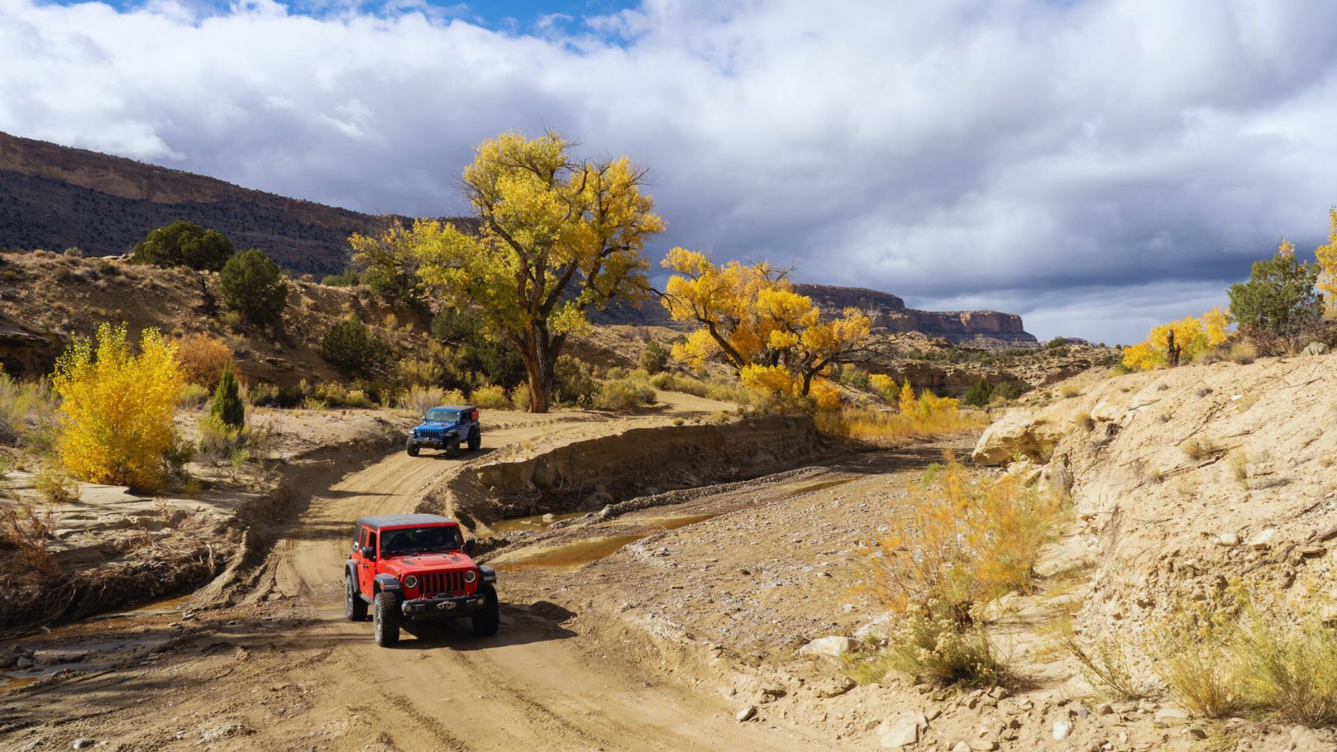 Two off-road vehicles drive on a dirt path through a desert landscape. Autumn trees with golden leaves line the path under a cloudy blue sky.