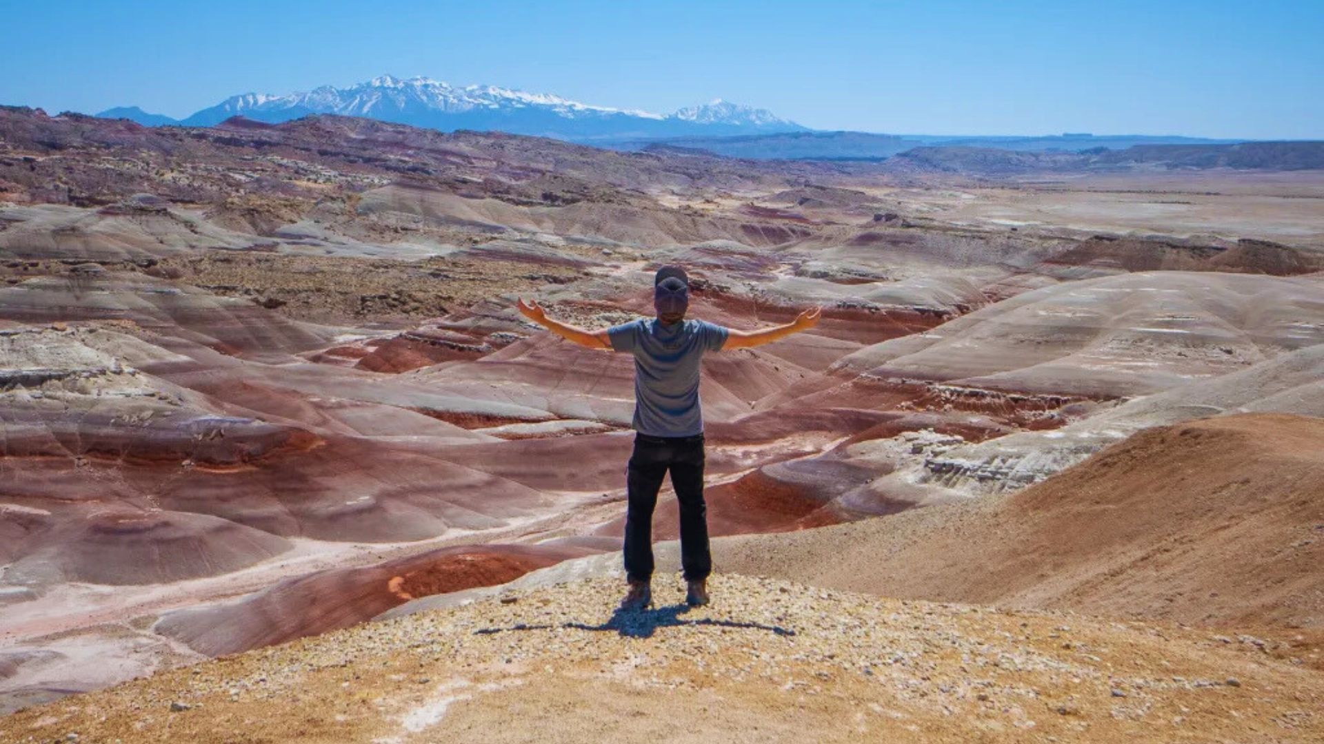 Person standing with arms outstretched on a hill, overlooking a vast, colorful desert landscape with layered red and brown earth, under a clear blue sky.