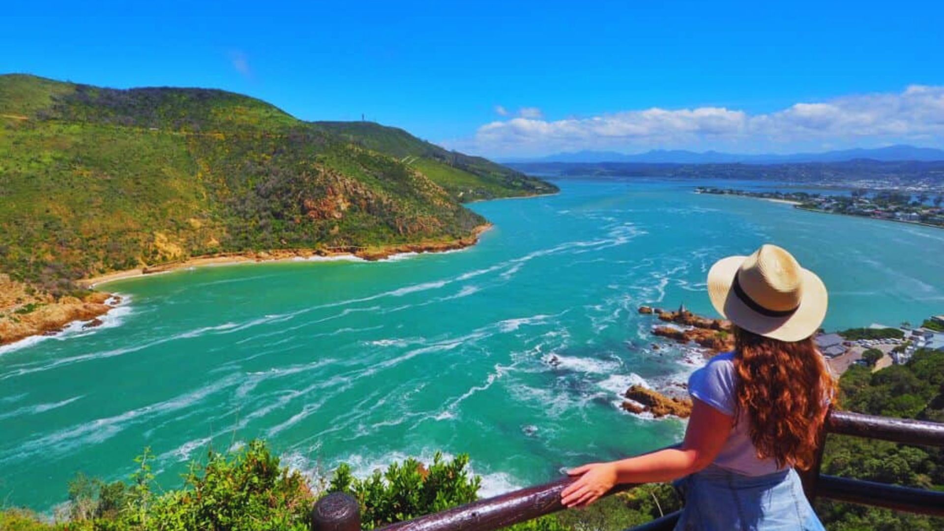A woman in a hat gazes at the ocean and mountains, enjoying the scenic view before her.