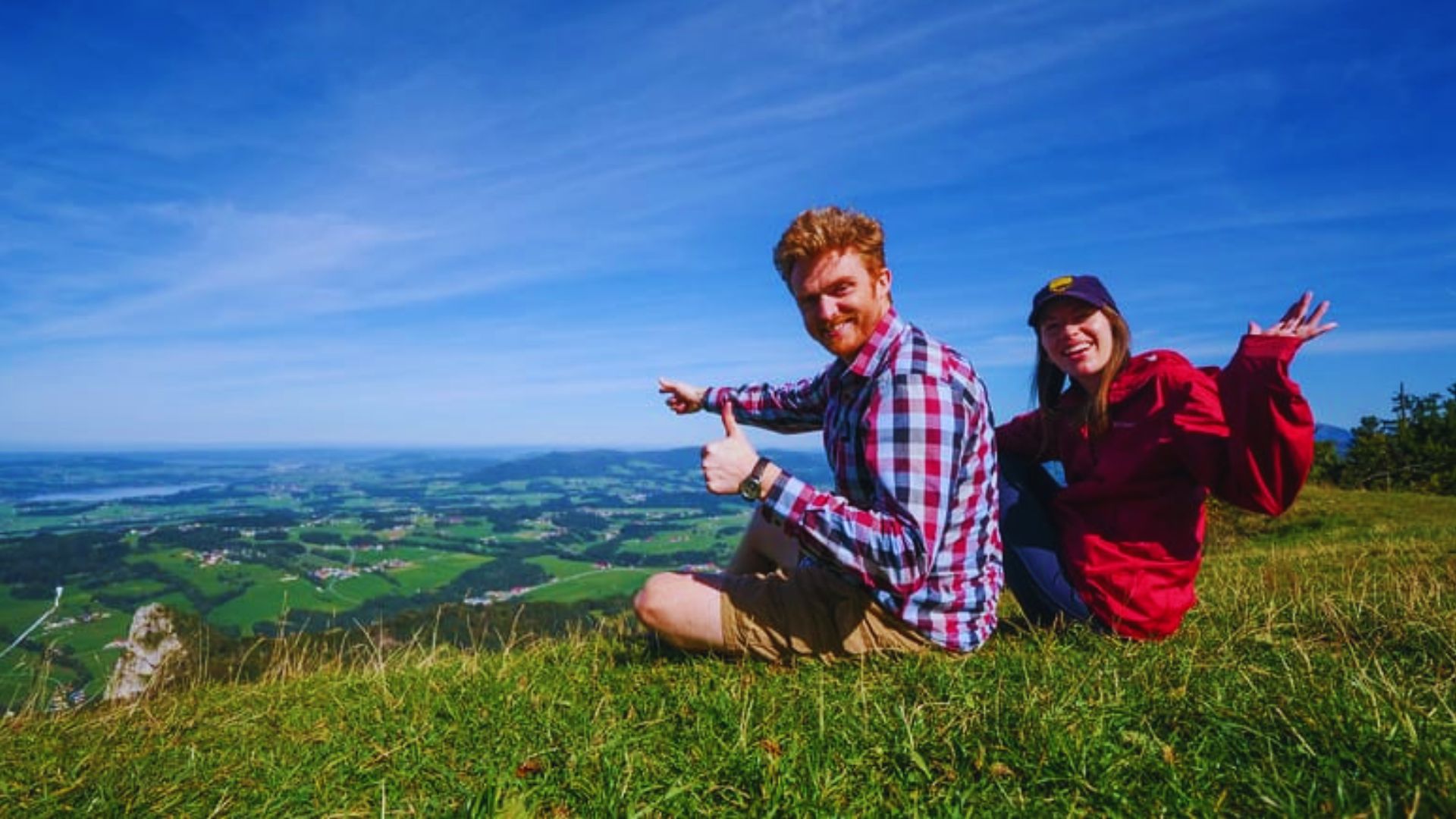 Two people sitting on a hilltop, smiling and giving thumbs up, enjoying a scenic view together.