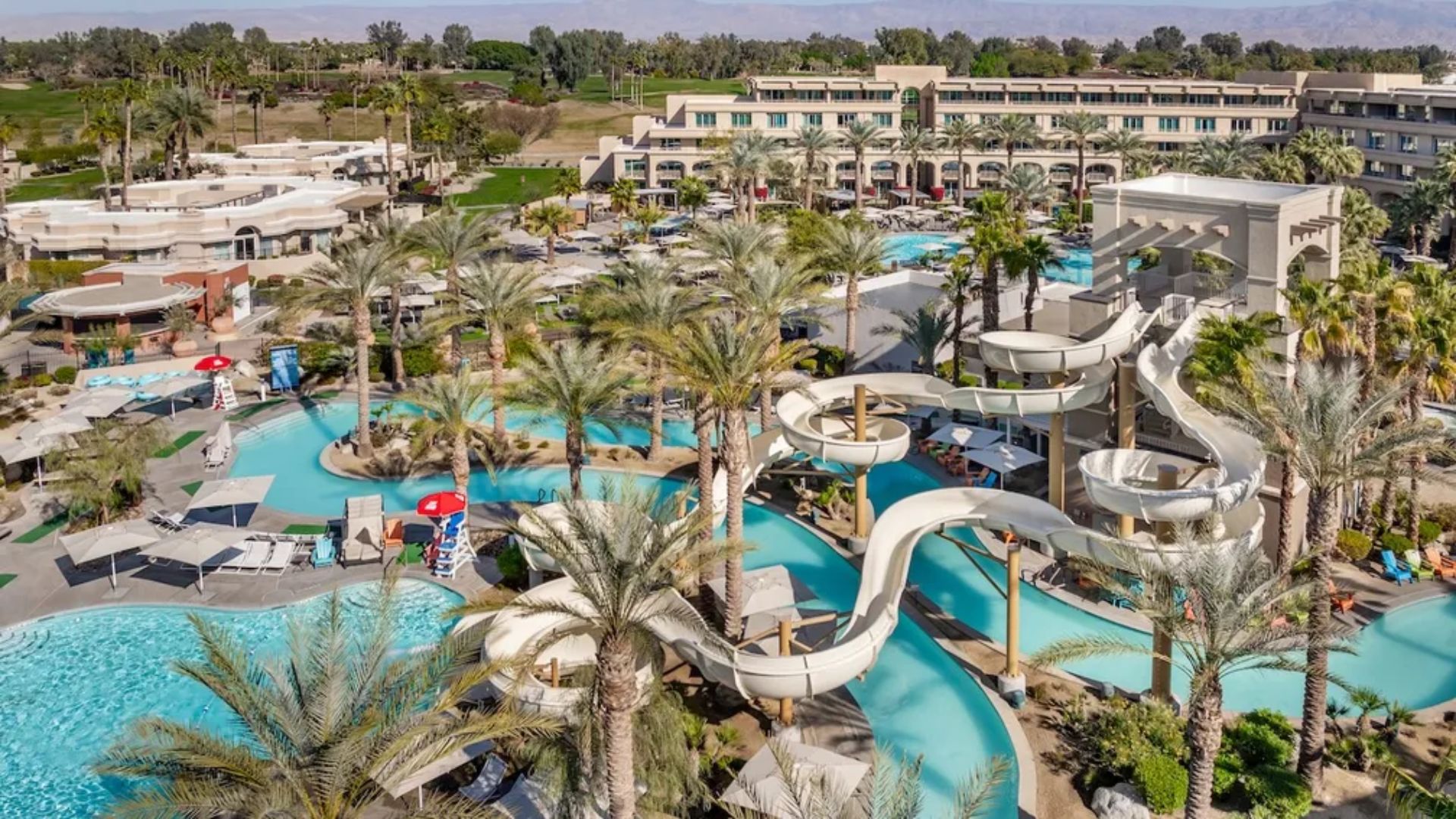 A large resort pool featuring multiple water slides, surrounded by lounge chairs and tropical landscaping.
